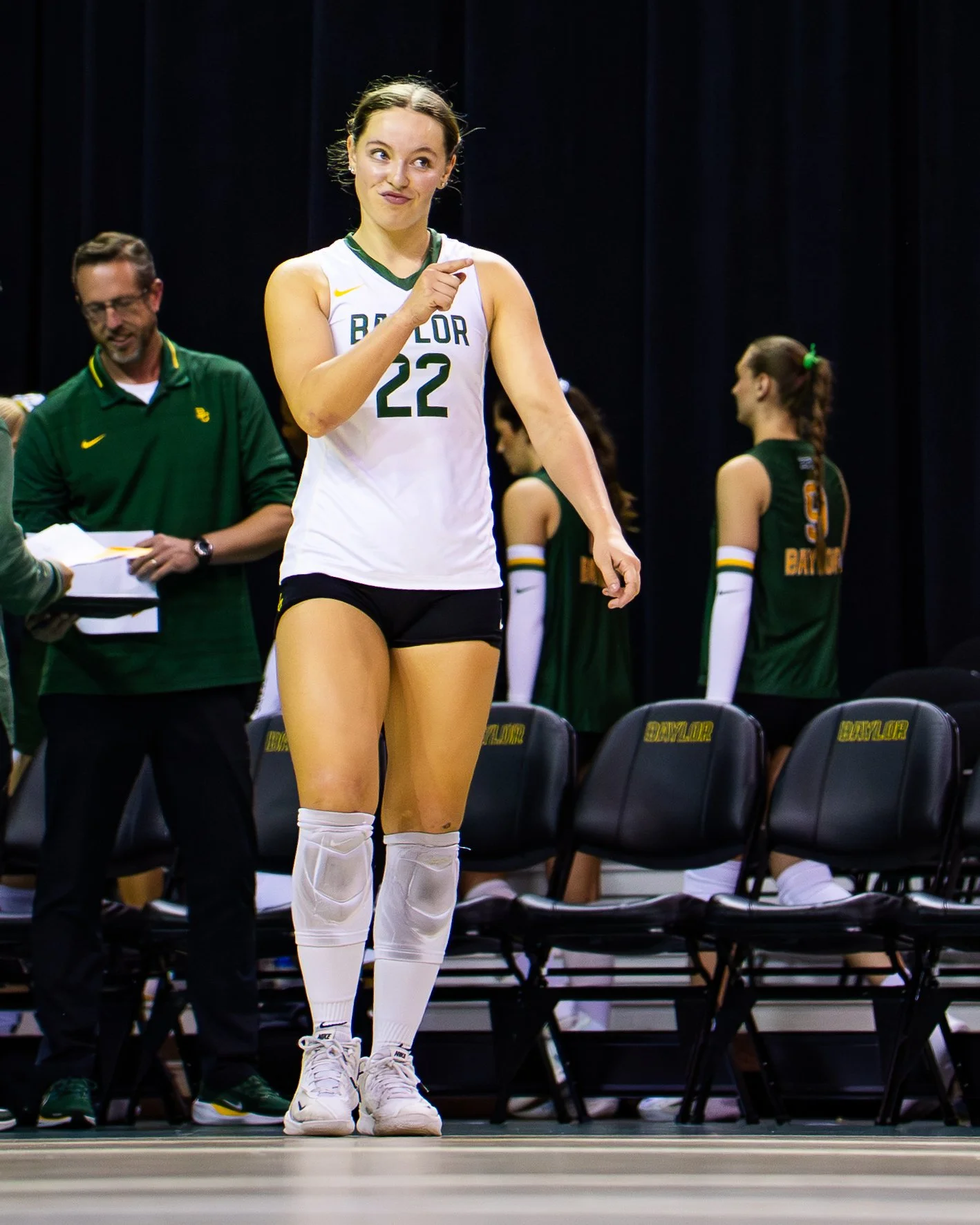 A female volleyball player wearing a white jersey with the number 22, white knee-high socks, and white athletic shoes standing on a court. She is pointing upward with her right hand, with a playful expression. In the background, other team members an
