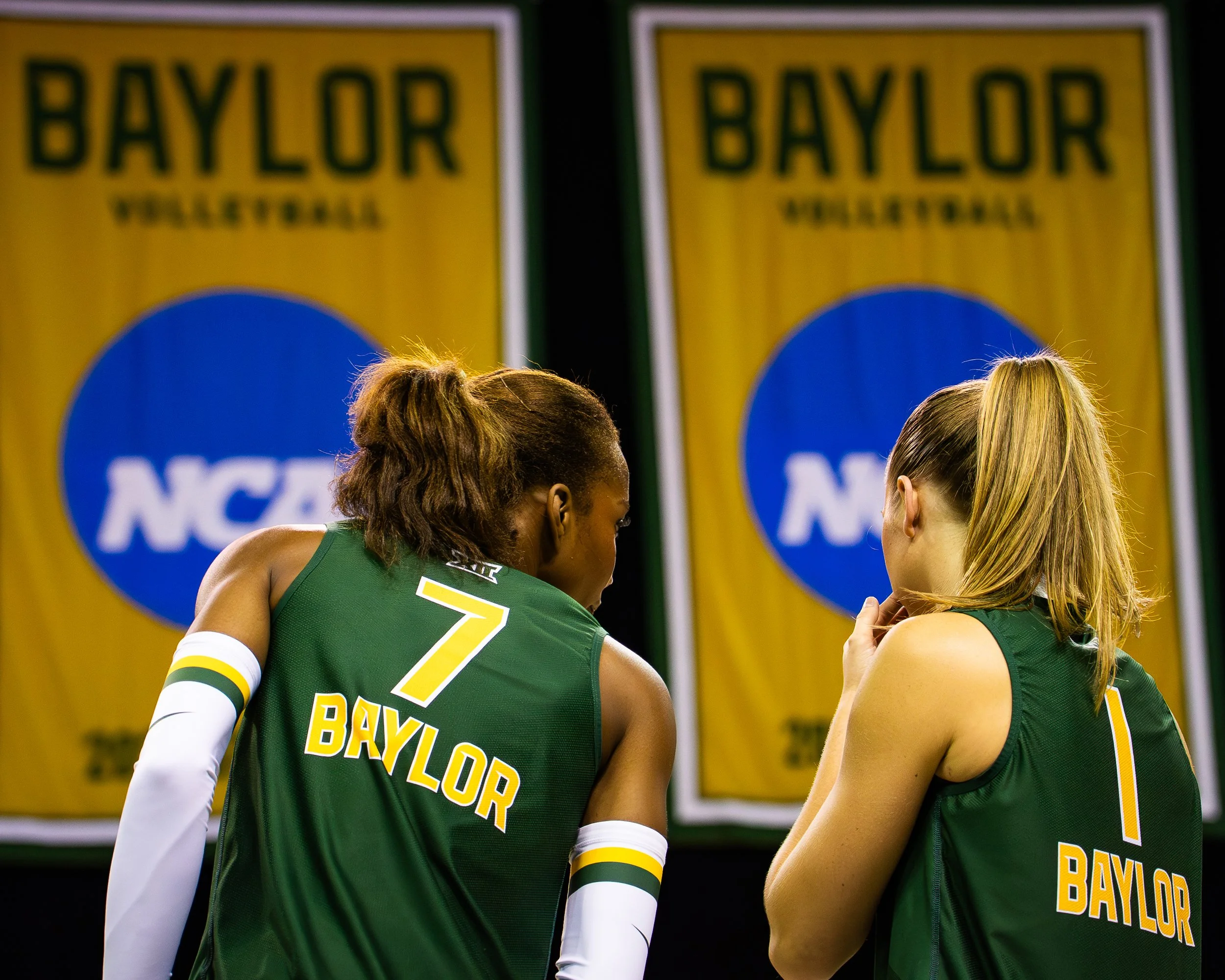 Two female volleyball players in green jerseys having a conversation during a match, with a yellow Baylor University banner in the background.