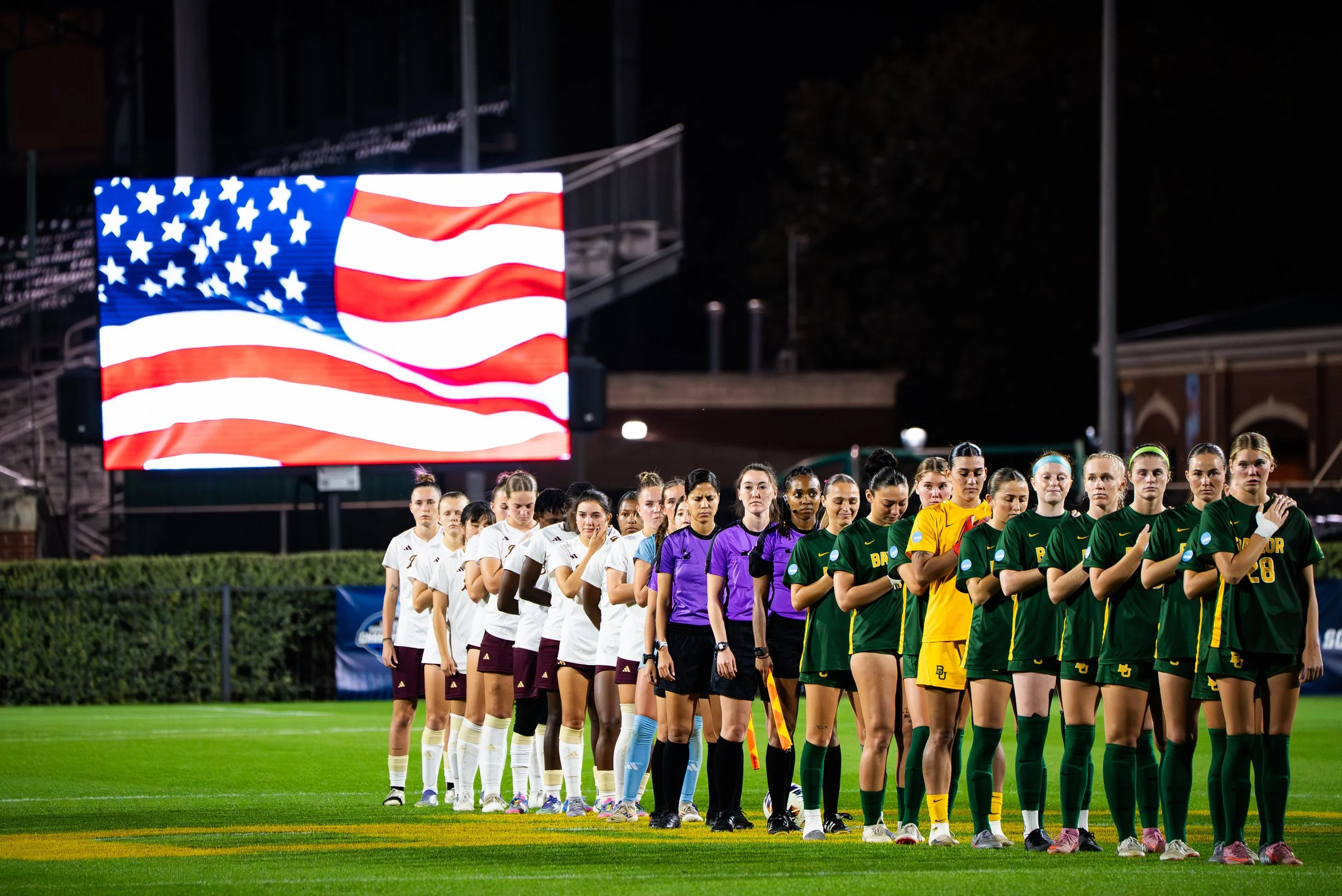 Women’s soccer players from different teams standing in a line on the field, A large American flag displayed on a screen behind them, under stadium lights at night.