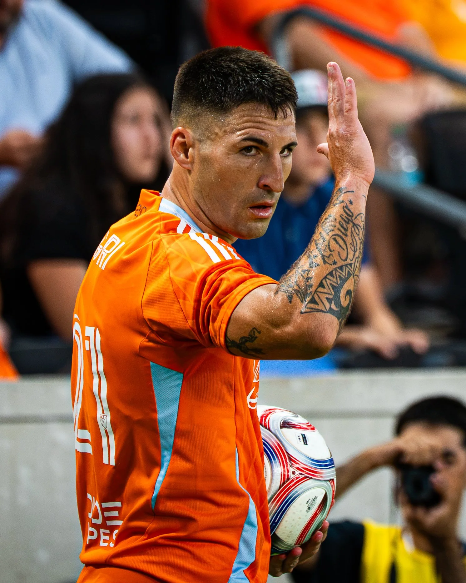 A male soccer player with short dark hair and tattoos on his left arm, wearing an orange and turquoise jersey, is holding a soccer ball and looking back over his shoulder.