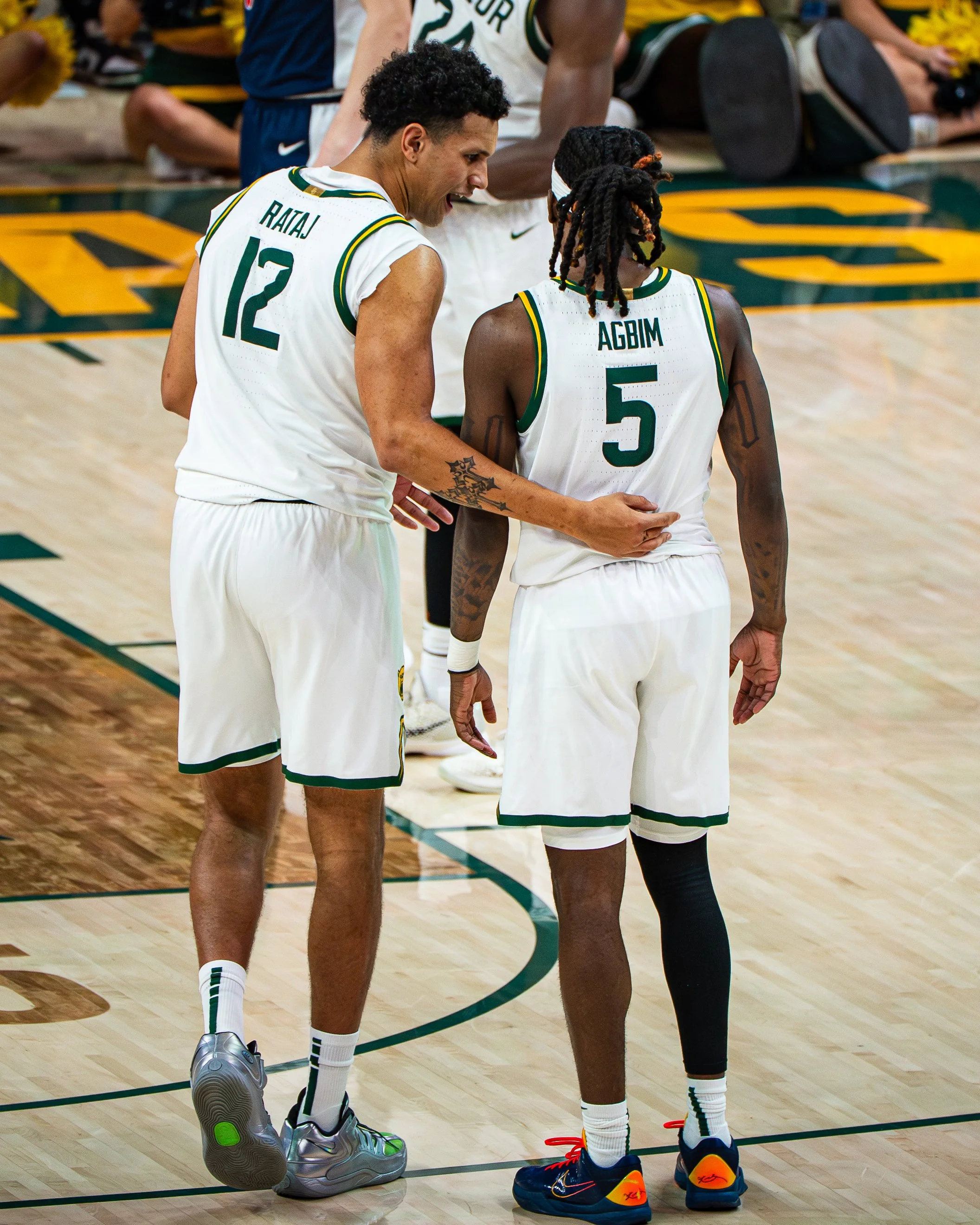 Two basketball players, one with a jersey labeled 'RATAJ 12' and the other with 'AGBIM 5', are standing close together on a basketball court, engaging in conversation. The player with the jersey 'RATAJ' has a tattoo on his right arm and is wearing si