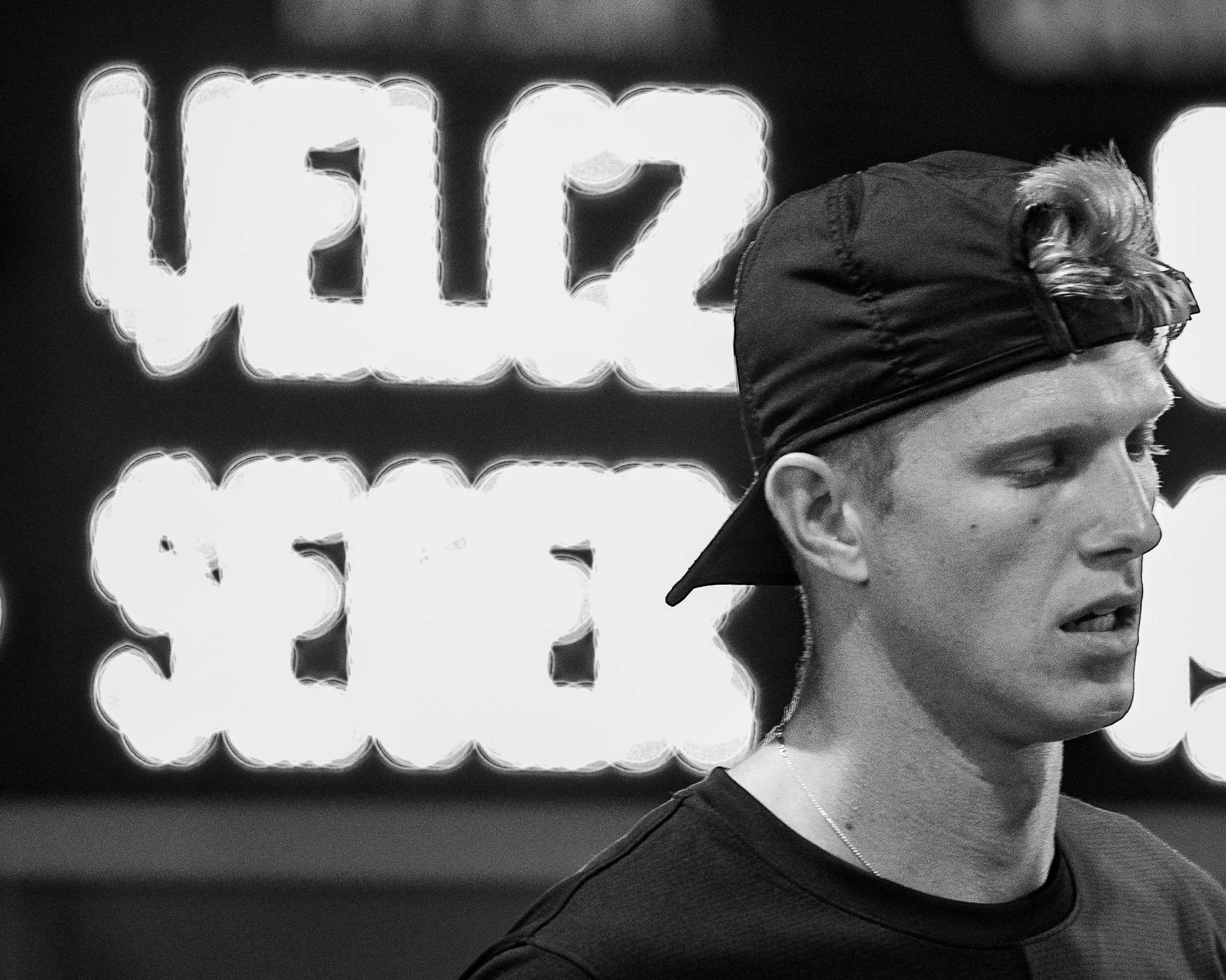 Close-up profile of a man wearing a black baseball cap backwards, in front of illuminated sign with large glowing letters.