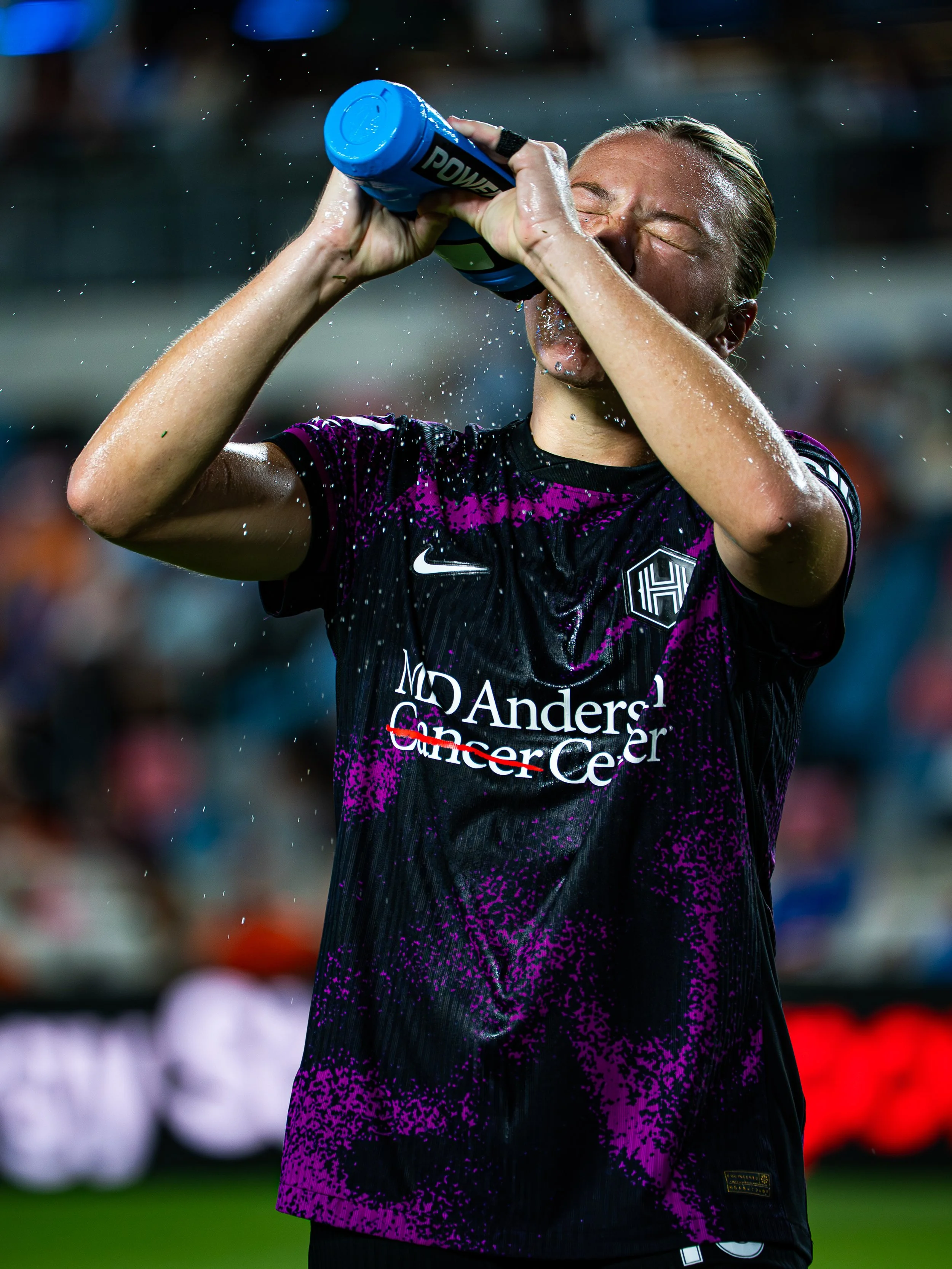 A female athlete with wet hair drinking from a blue water bottle during a game. She is wearing a black and purple sports jersey with 'ND Anderson Cancer Center' printed on it.