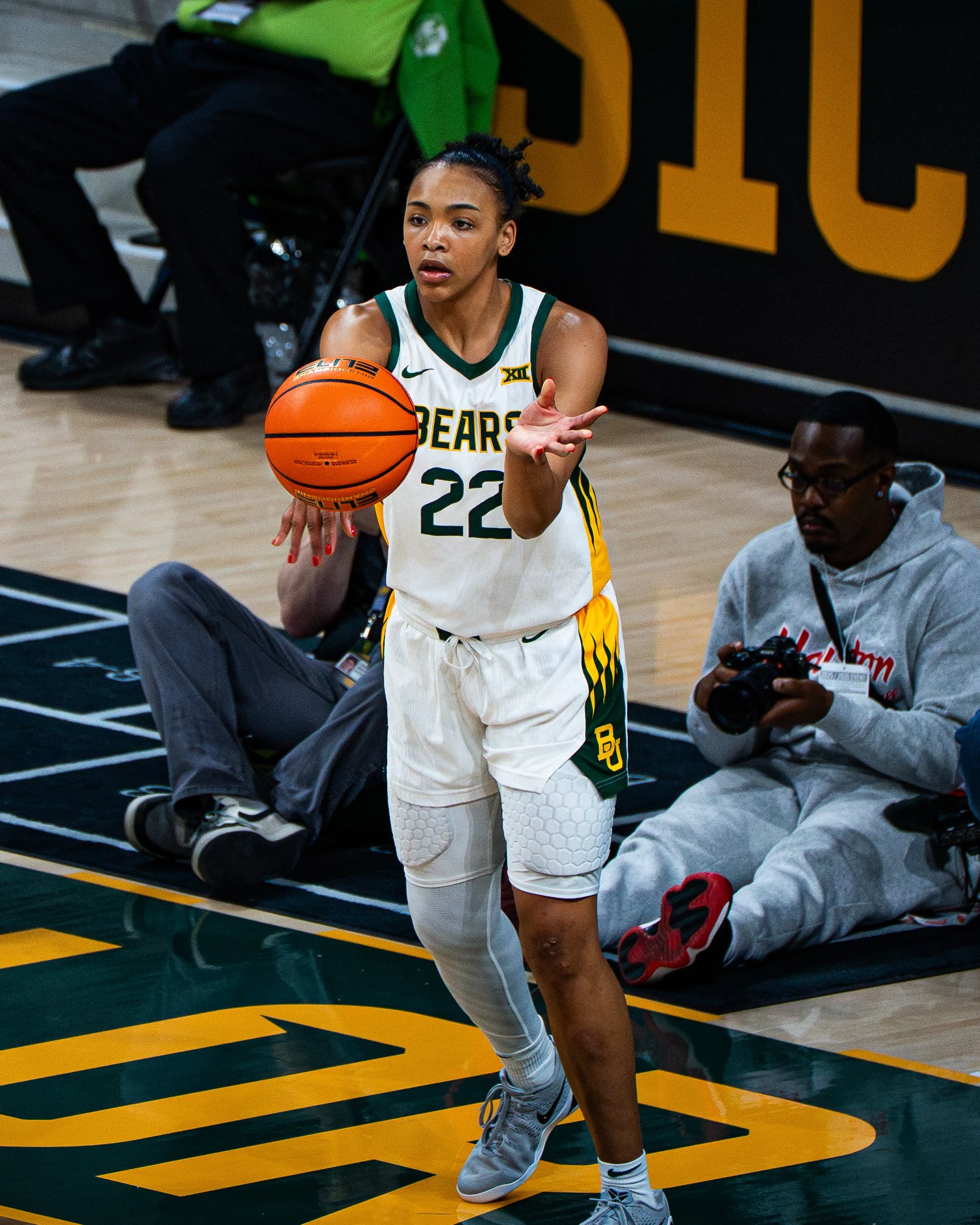 A female basketball player in a white and green uniform with number 22 is holding a basketball on a basketball court. She has her hair in a bun, appears focused, and is wearing white knee pads and sneakers. A man in glasses and a gray hoodie is sitti