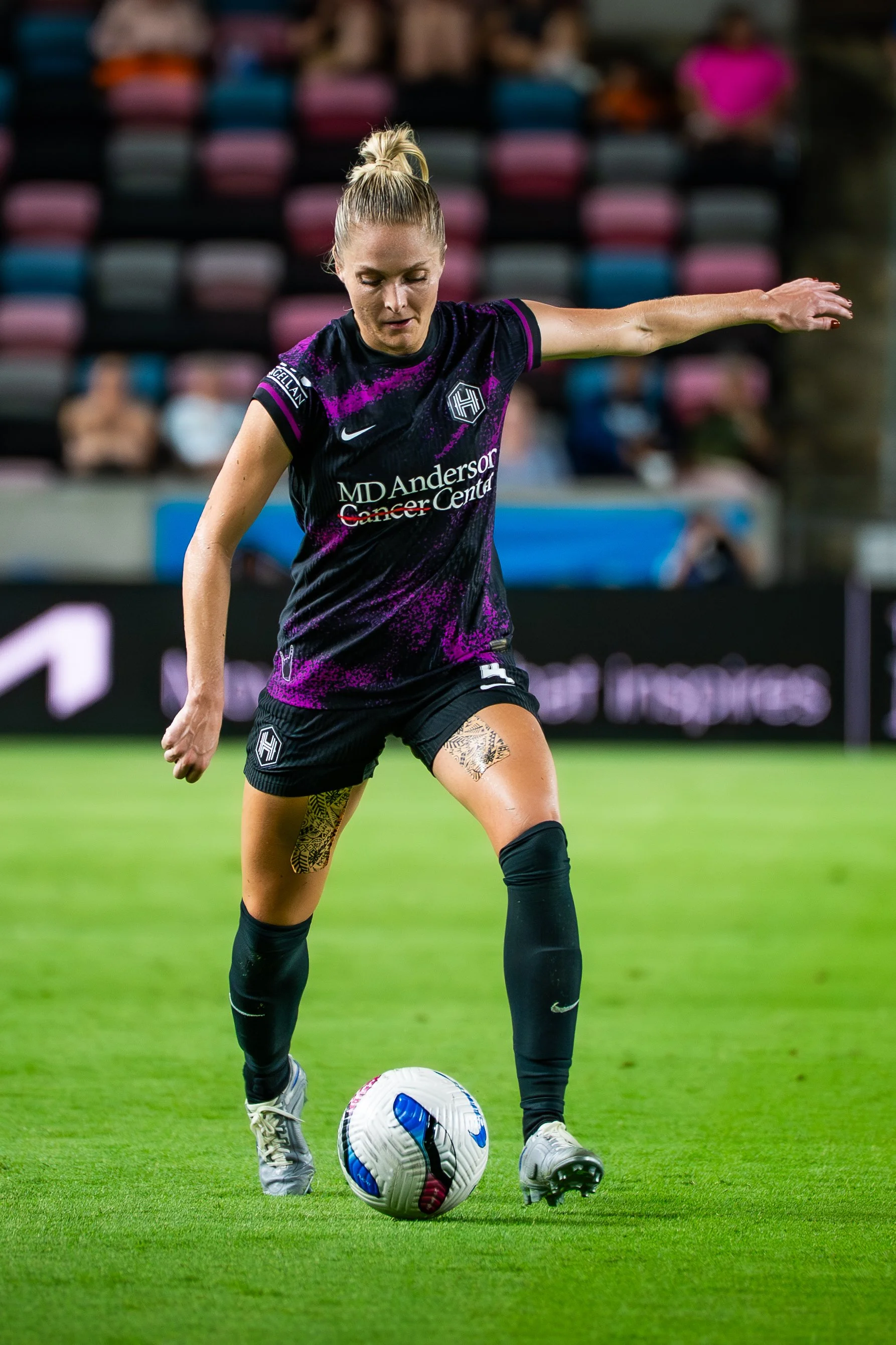 A female soccer player in a black and purple uniform kicking a white and blue soccer ball on the field, with colorful stadium seats in the background.
