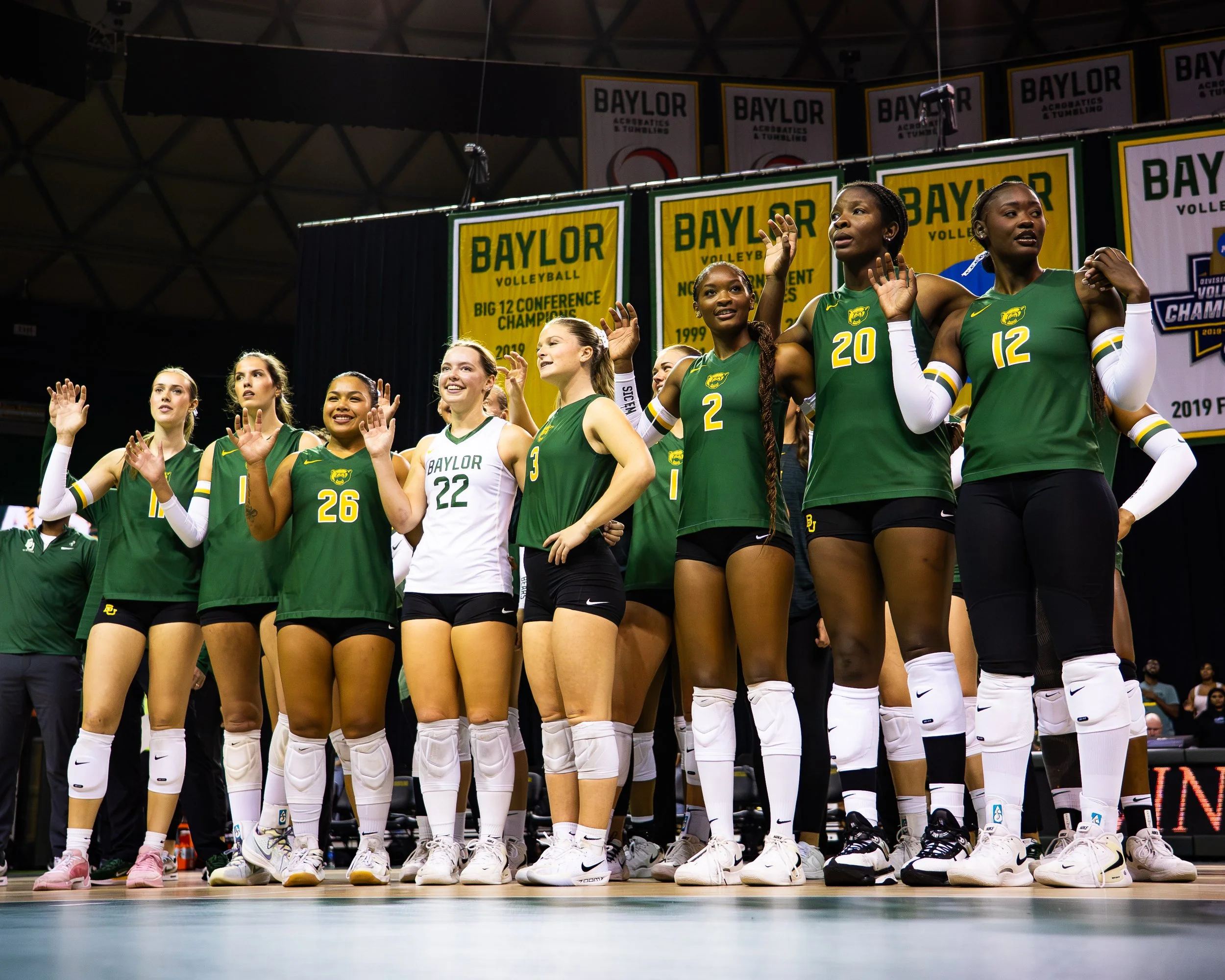 A group of female volleyball players standing in a line on a volleyball court, wearing green jerseys with yellow and white accents, and white knee pads, with an indoor sports arena background and banners that read 'Baylor Volleyball Big 12 Conference