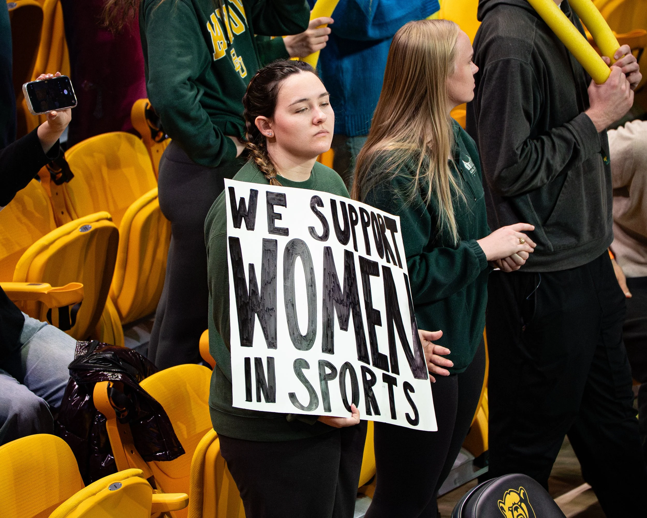 A woman holding a protest sign at a sporting event. She is wearing a green sweatshirt with a braid over her shoulder. The sign reads 'We support YOU in sports.' There are other people standing nearby, some holding yellow foam sticks. The background s