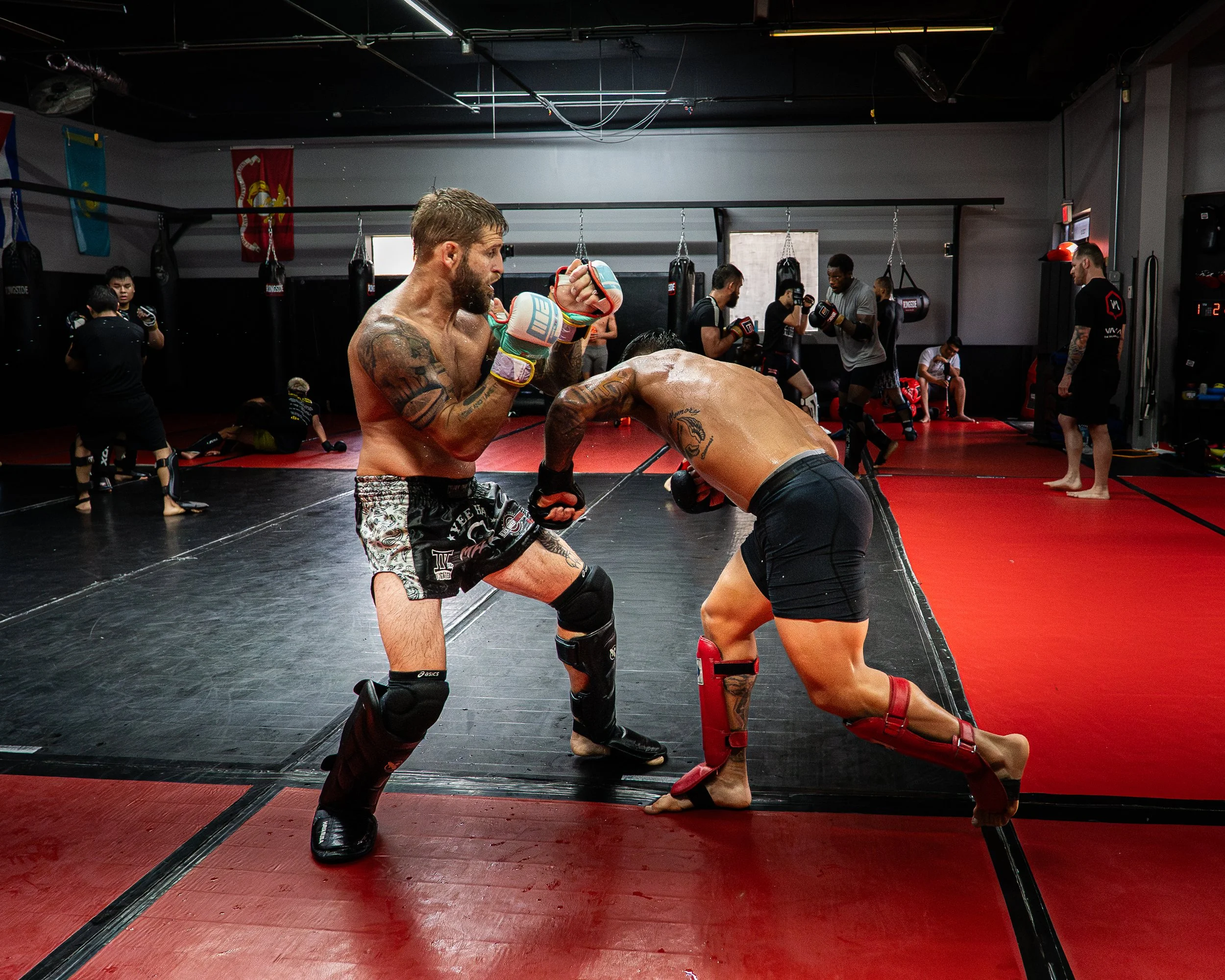 A mixed martial arts fight taking place in a gym with several spectators and fighters in the background. The fighters are using boxing gloves and protective gear, engaged in close combat.