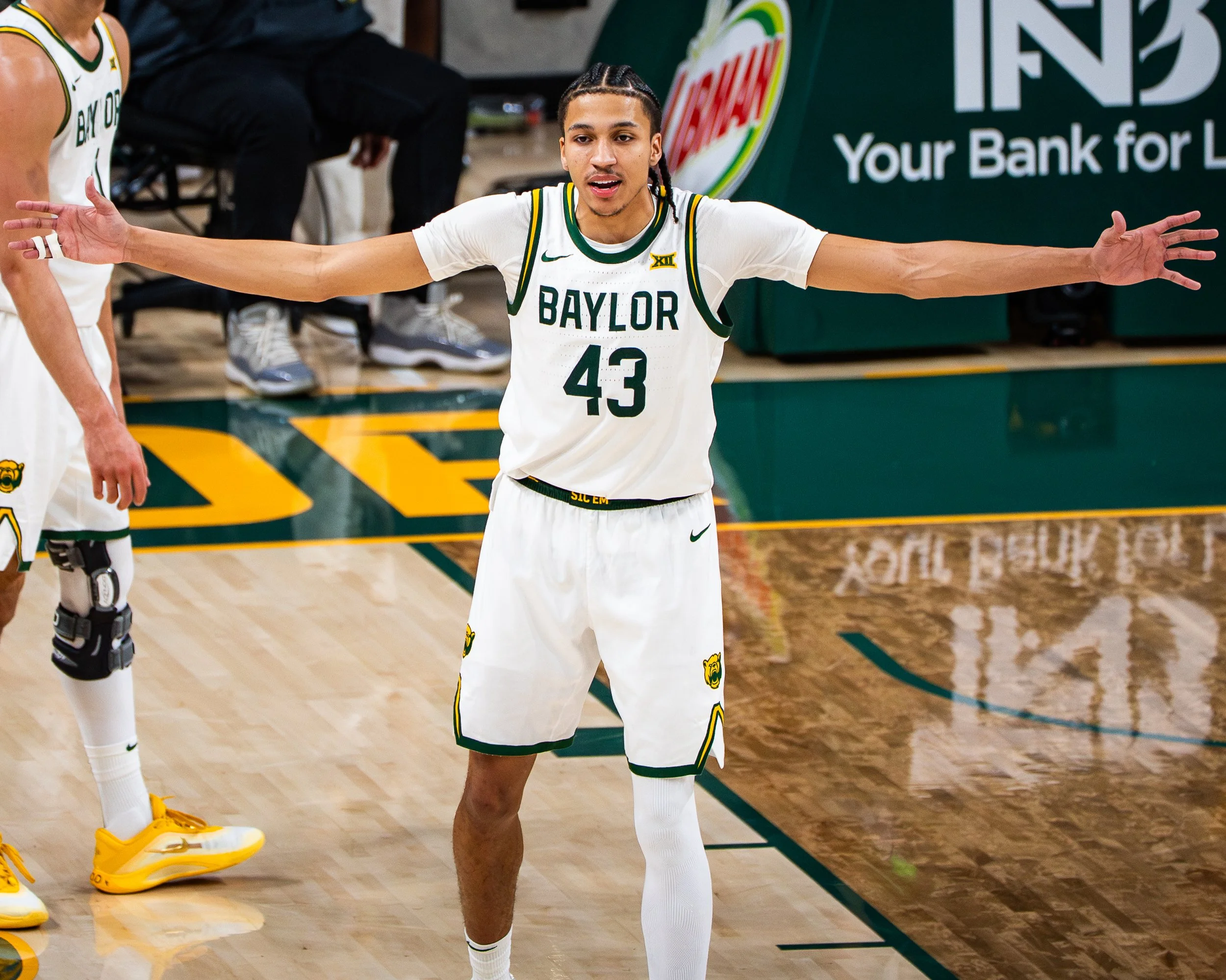A basketball player in a white Baylor University uniform with the number 43, celebrating on the court with arms outstretched. Part of another player in yellow and white uniform and a seated person in black are visible in the background.