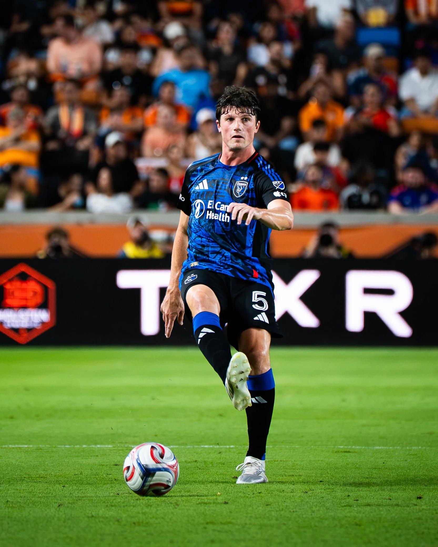 Soccer player in black and blue uniform running on field with a soccer ball at his feet at night with a crowd in the background