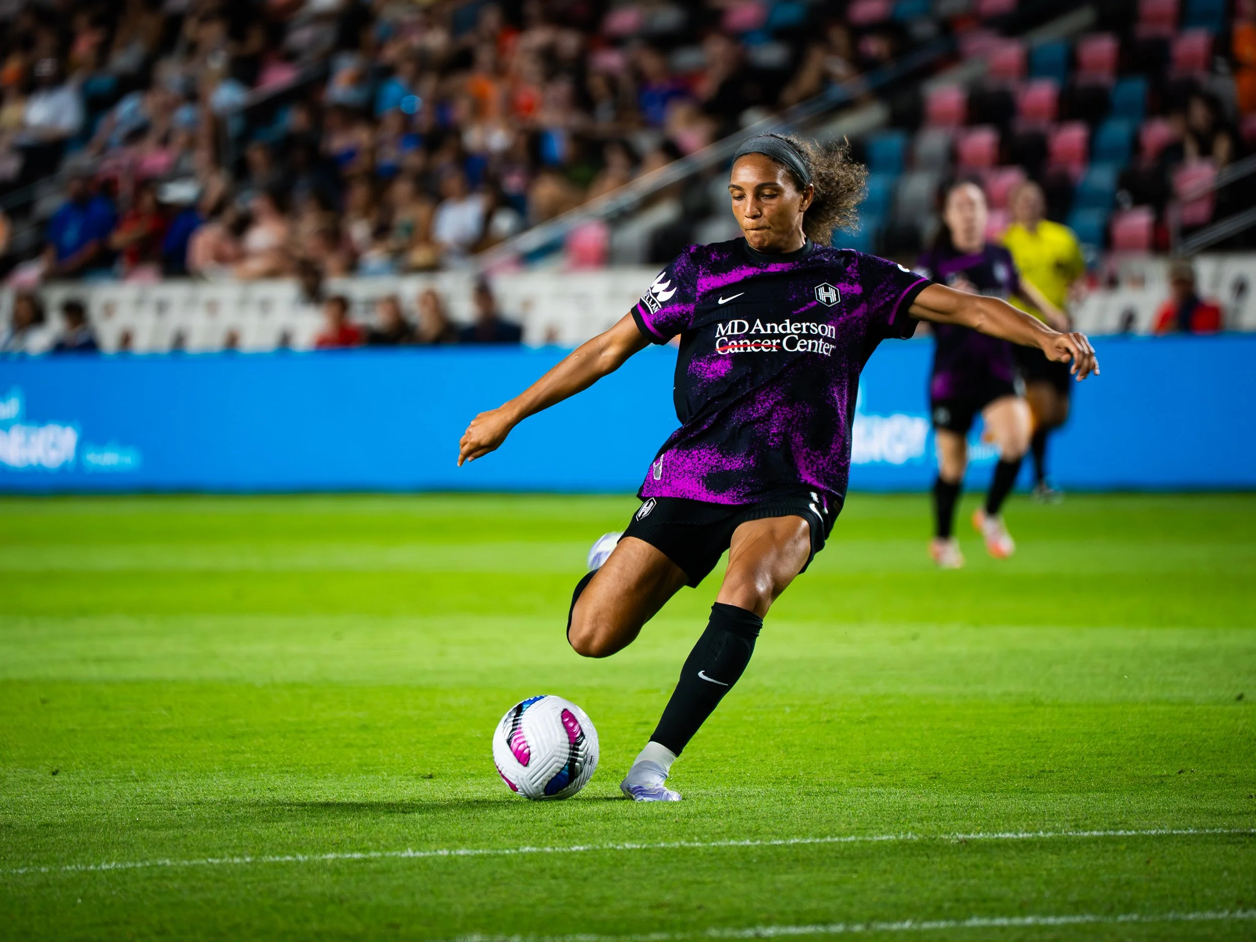 Female soccer player in black and purple uniform kicking a soccer ball on a field during a match with stadium spectators in the background.