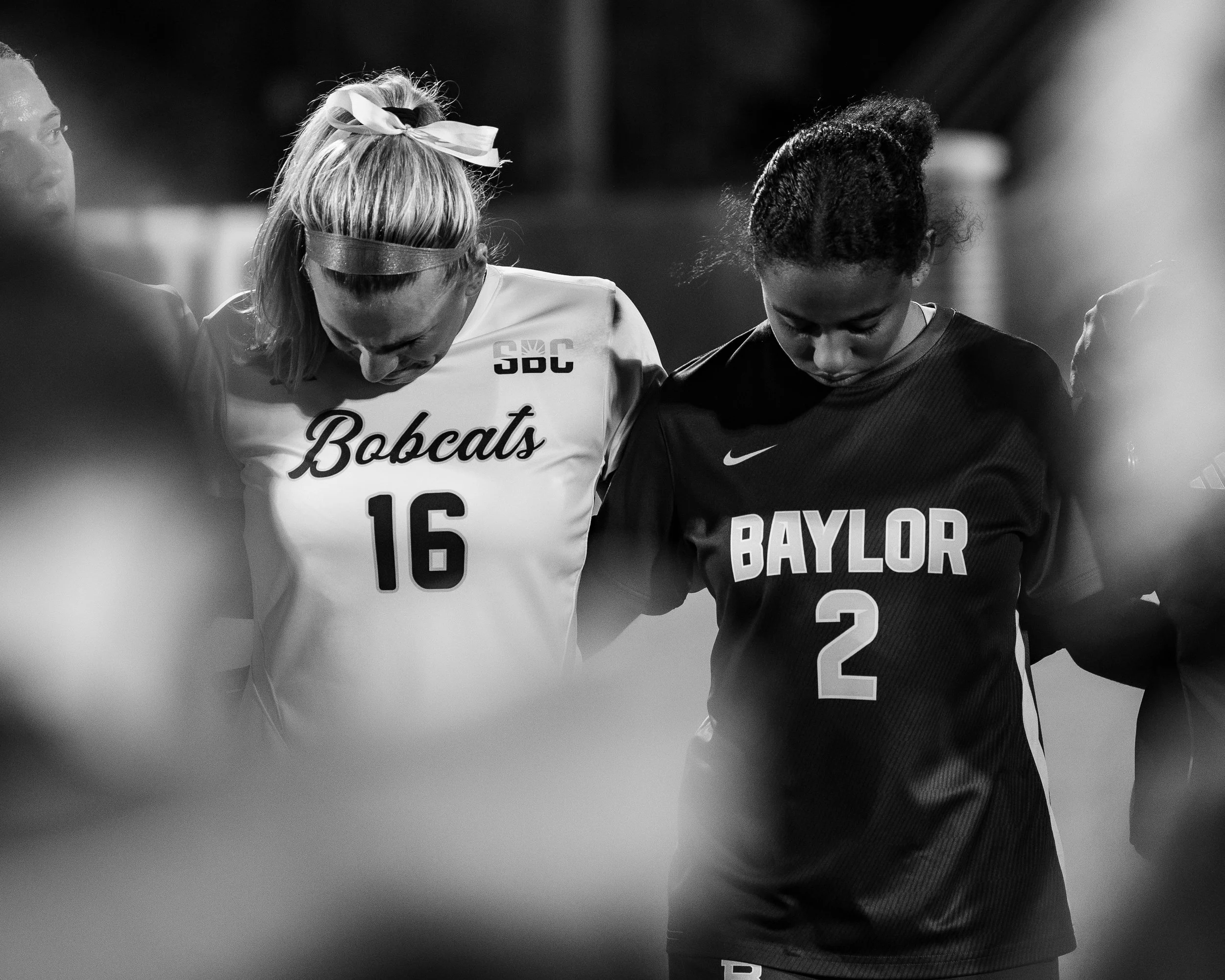Three female athletes with heads bowed and eyes closed, wearing sports uniforms with visible team names and numbers, standing together in a moment of reflection or prayer.