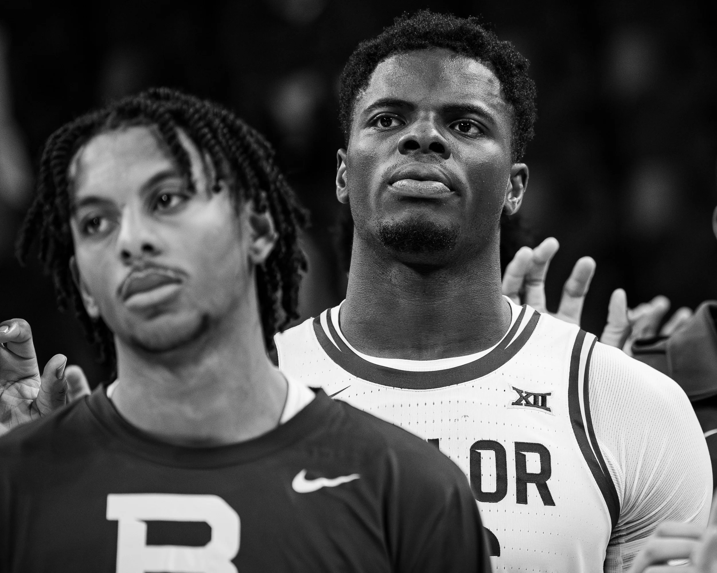 Two basketball players standing side by side; the player on the right is in a white jersey with the Big 12 Conference logo, making a gesture with his fingers, while the player on the left has dreadlocks and is looking slightly upward.