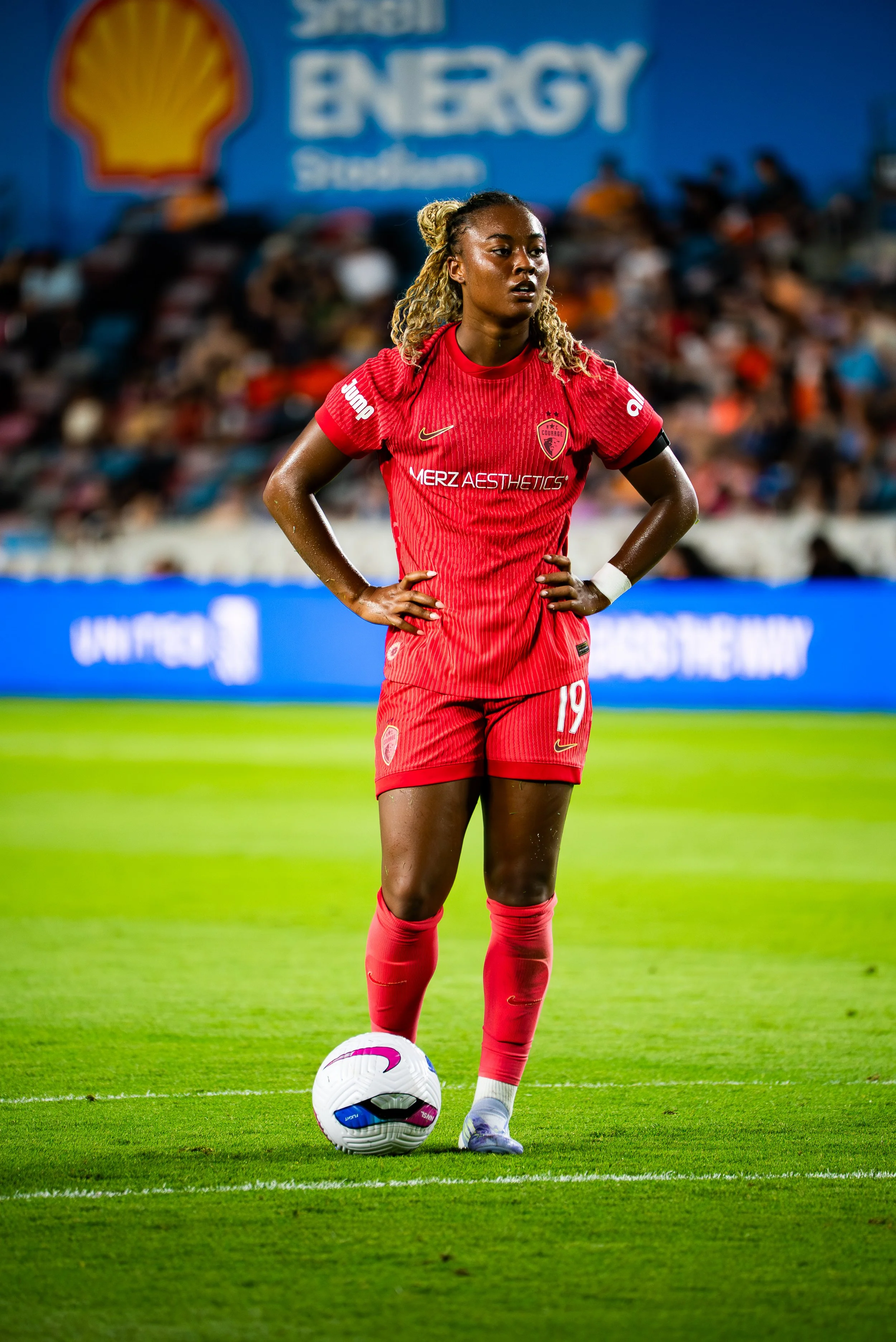 A female soccer player standing on the field with a soccer ball at her feet, wearing a red jersey and shorts, with a stadium and crowd in the background.