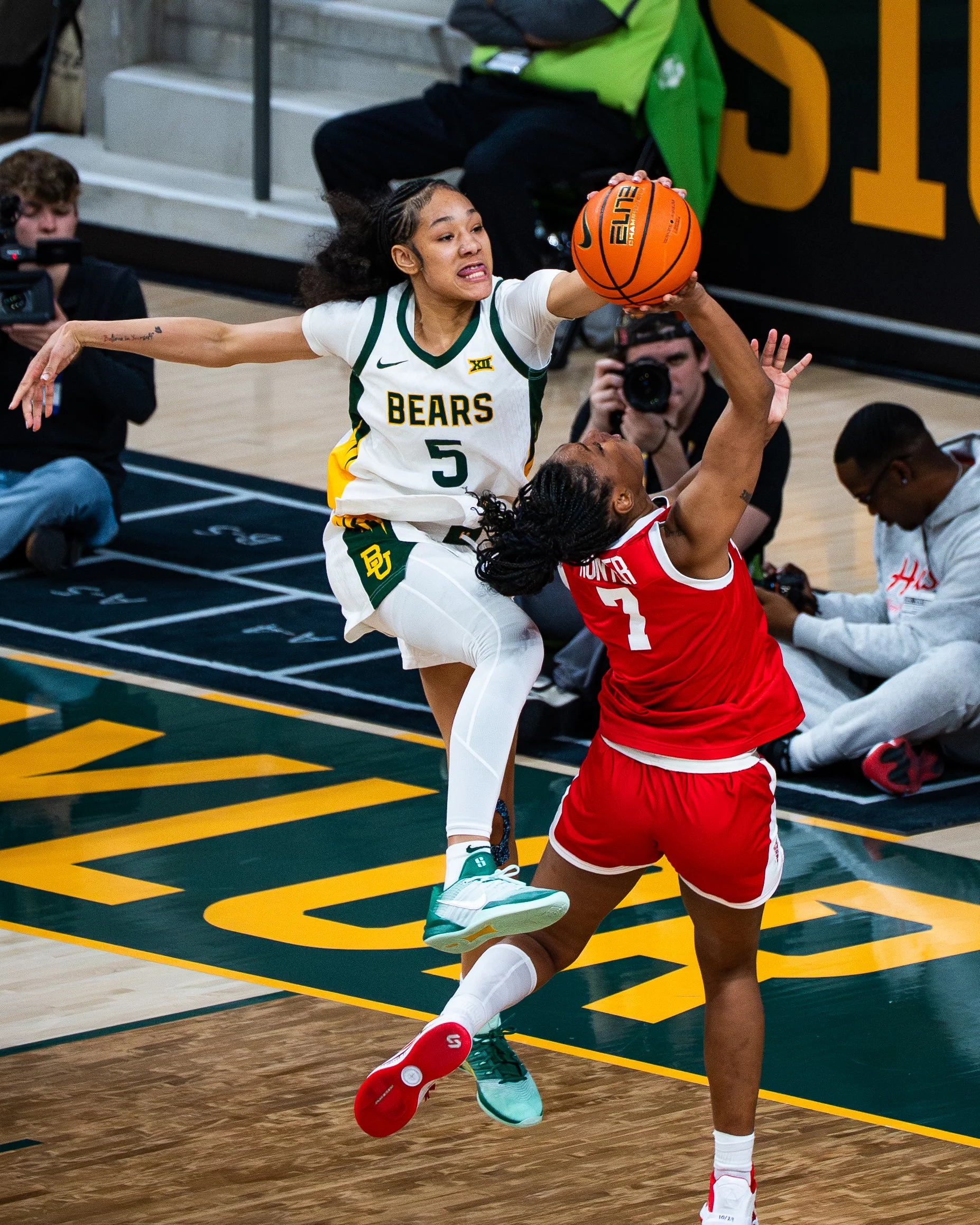 Two women playing basketball on an indoor court, with one attempting a shot and the other attempting to block. The woman in white with the number 5 jersey is jumping and reaching for the ball, while the woman in red is defending. Several photographer