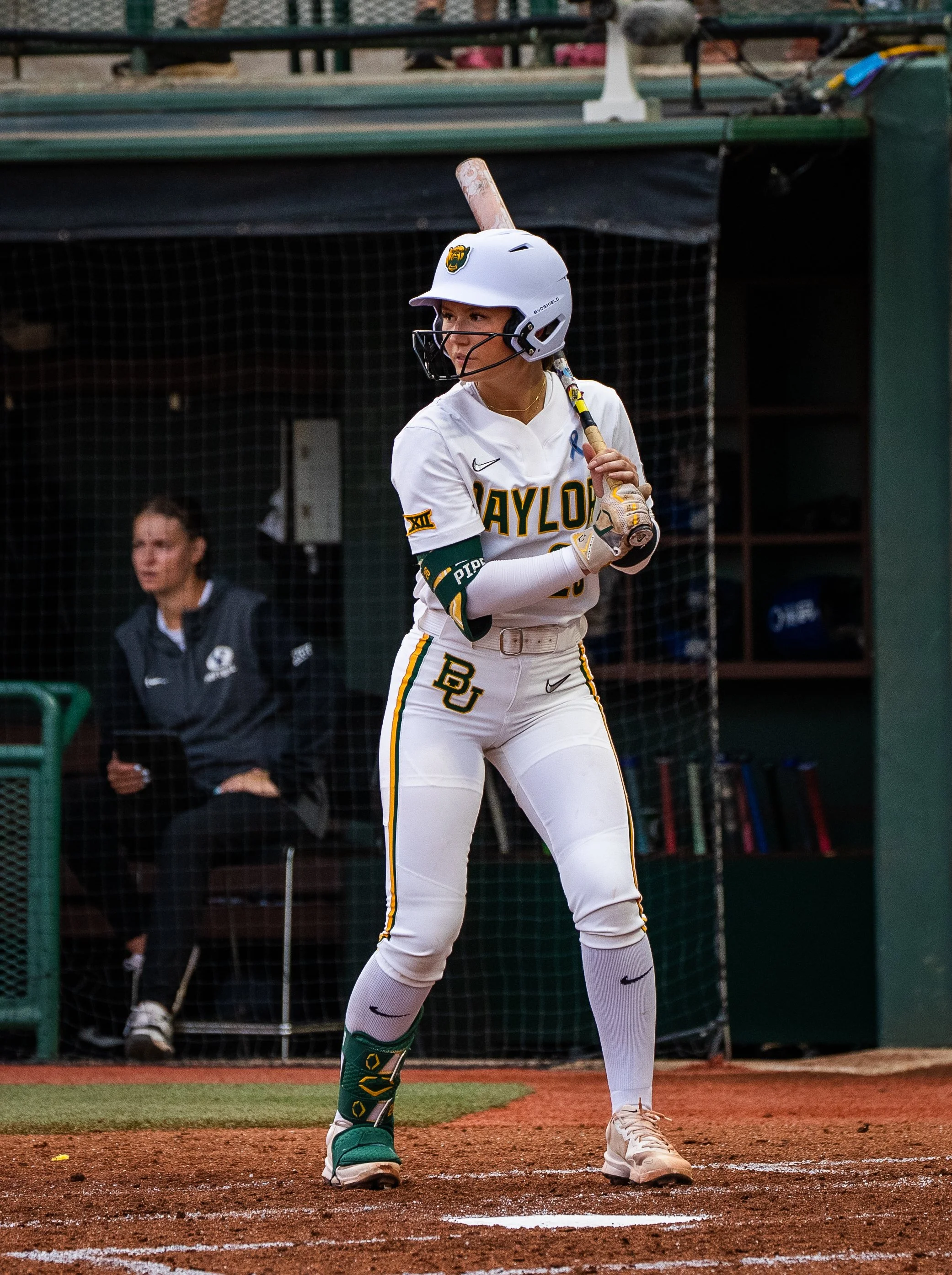 A female softball player wearing a white uniform with green and yellow accents, and a helmet, stands at home plate holding a bat. She is ready to bat on a softball field with a dugout, netting, and team members in the background.