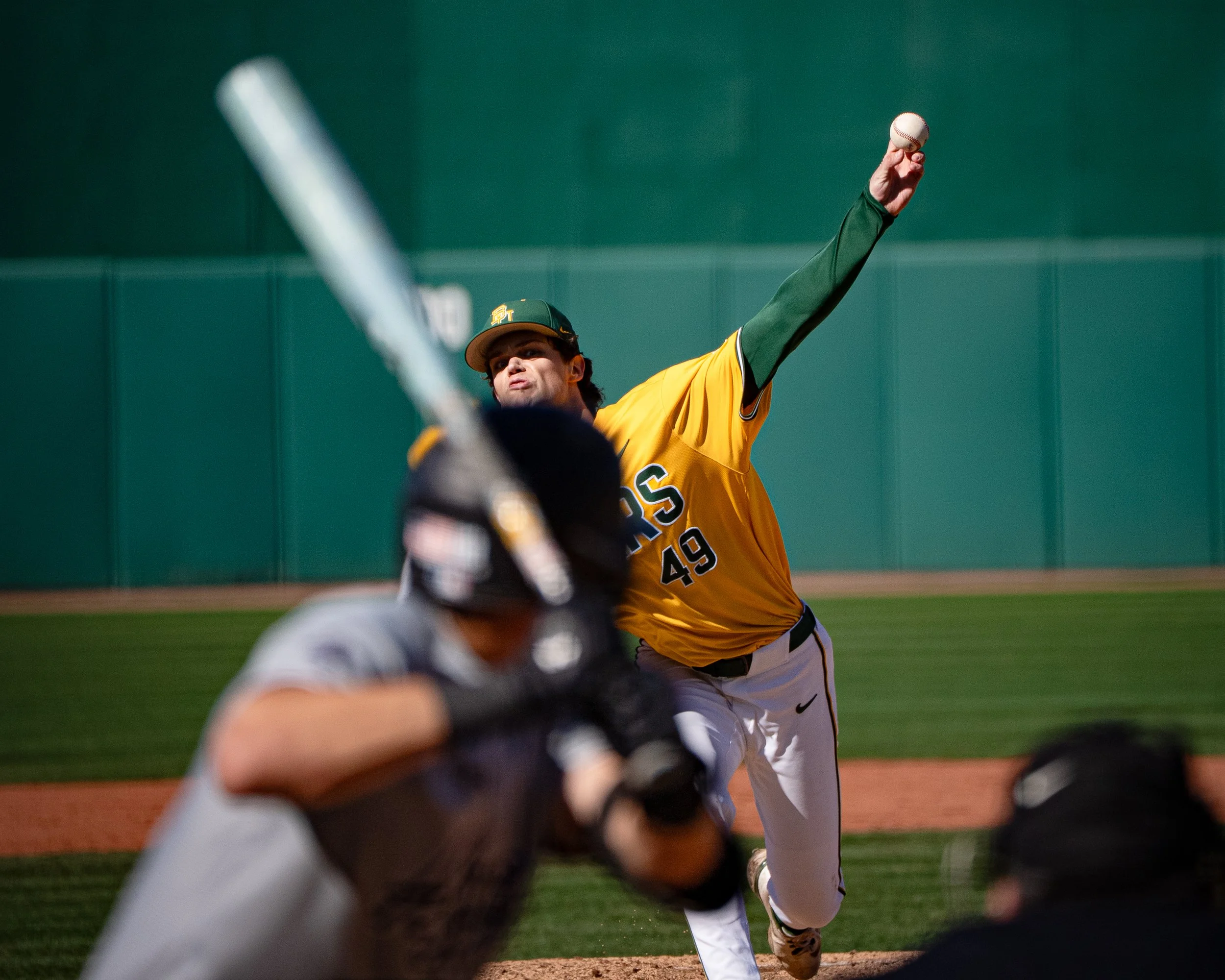 A baseball player in a yellow and green uniform throwing a pitch during a game, with other players in the foreground and a green wall in the background.