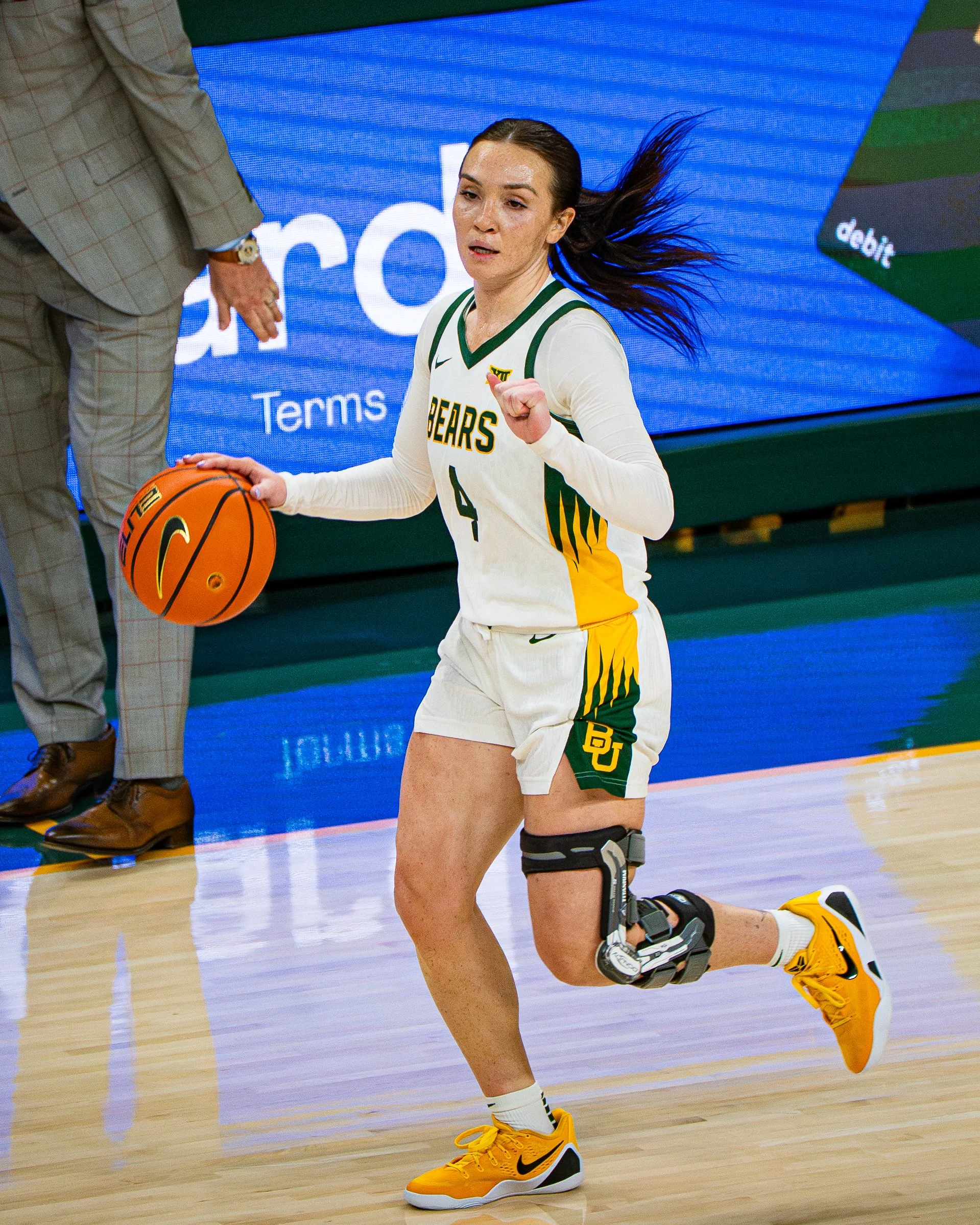 A female basketball player in a white and green uniform with yellow shoes is kneeling on the court during a game.
