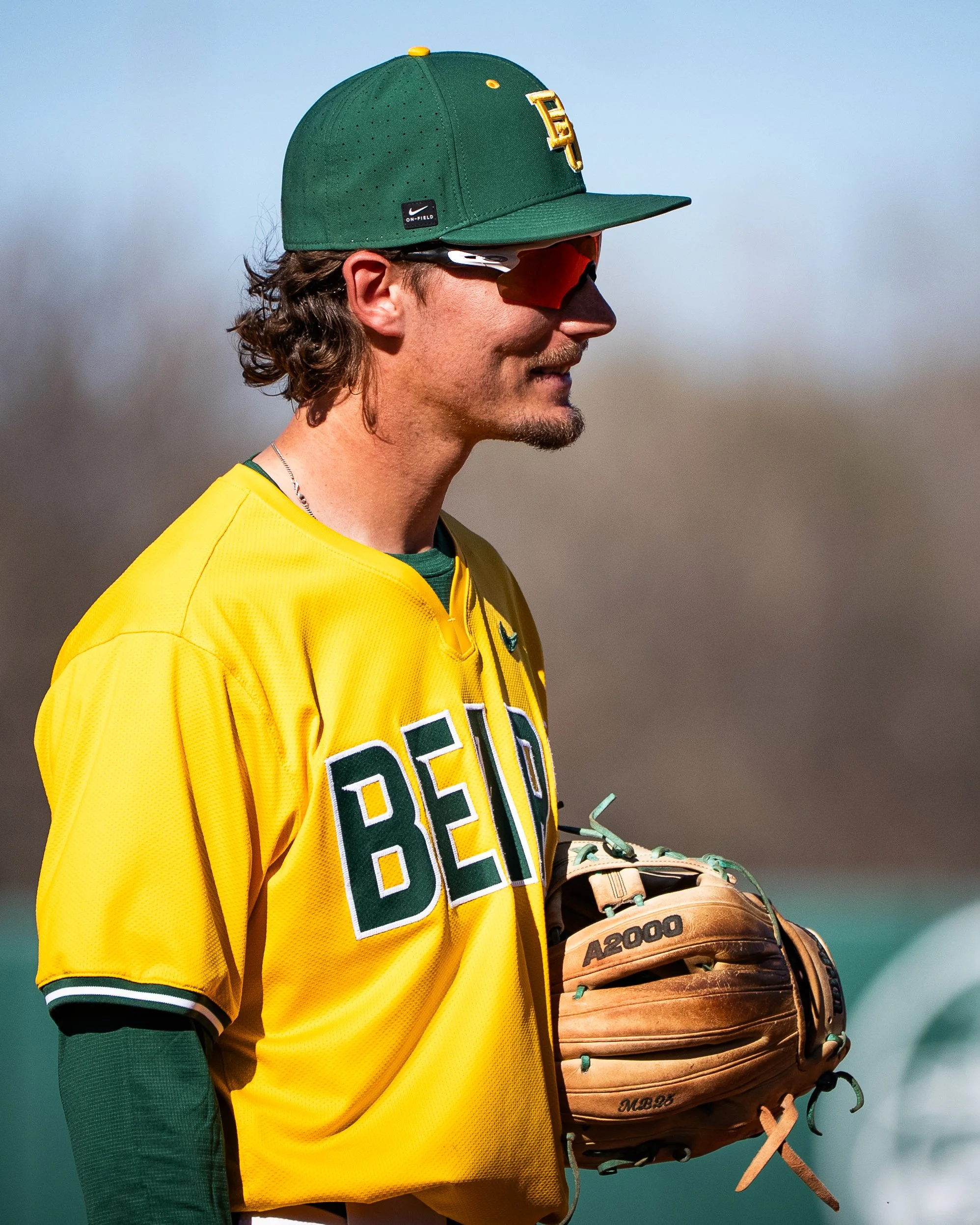 Close-up of a male baseball player wearing a green cap with yellow detail and a yellow jersey, holding a brown glove with green laces, standing outdoors under a blue sky.