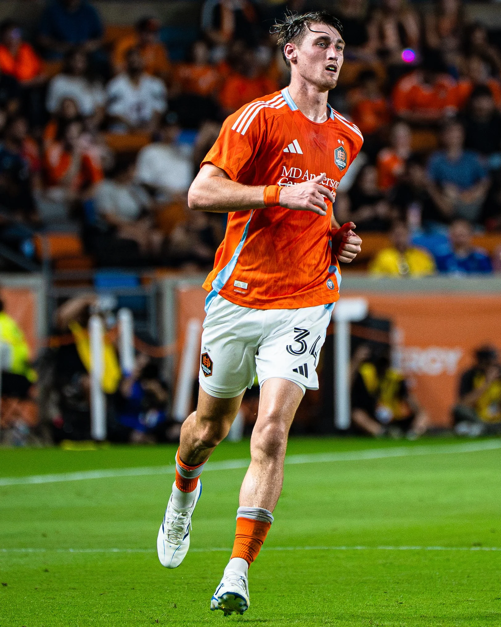 A male soccer player running on the field during a match, wearing an orange jersey and white shorts, with a crowd of spectators in the background.