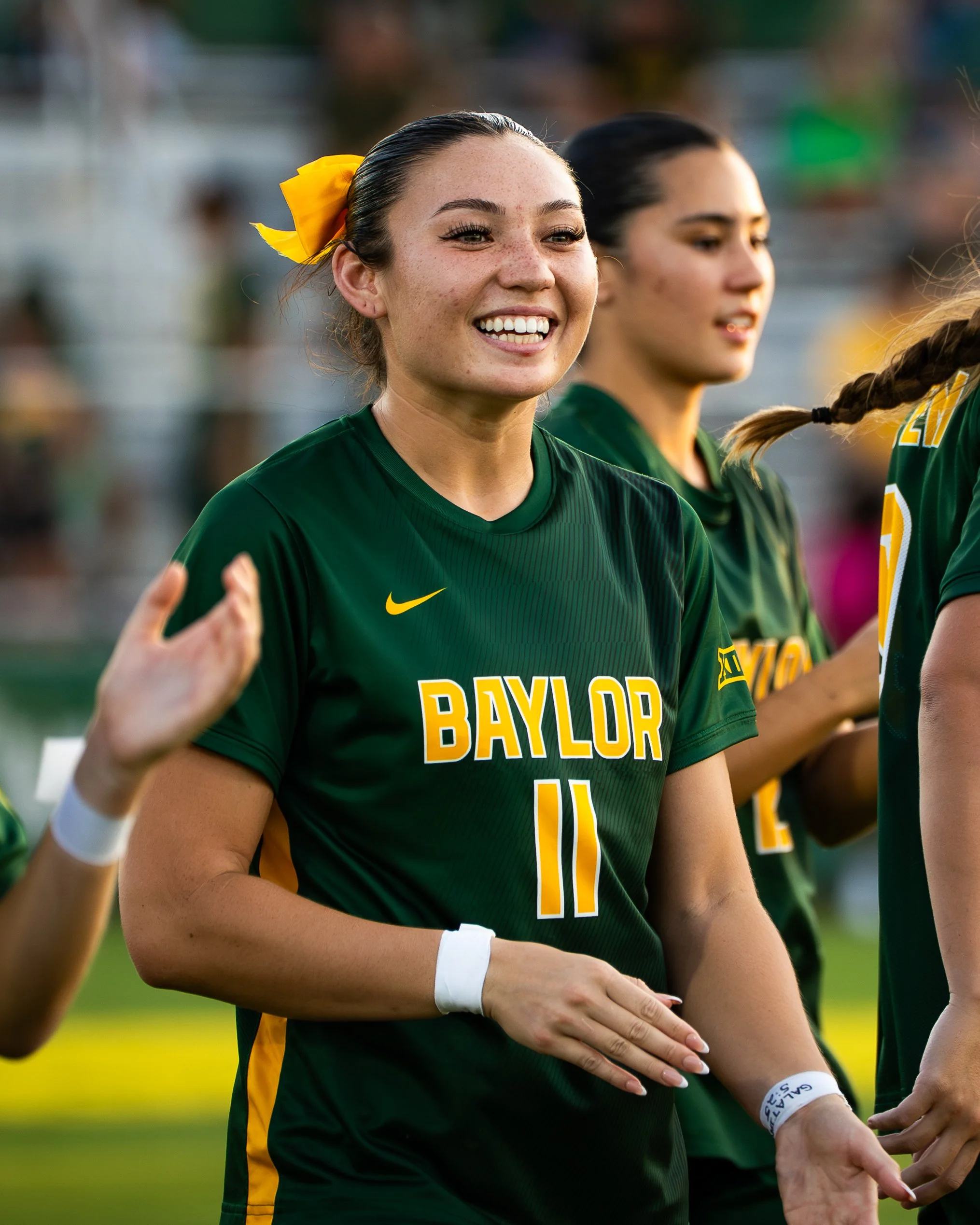 Close-up of a female soccer player with freckles smiling and wearing a green Baylor jersey with the number 11, with her teammates in the background.