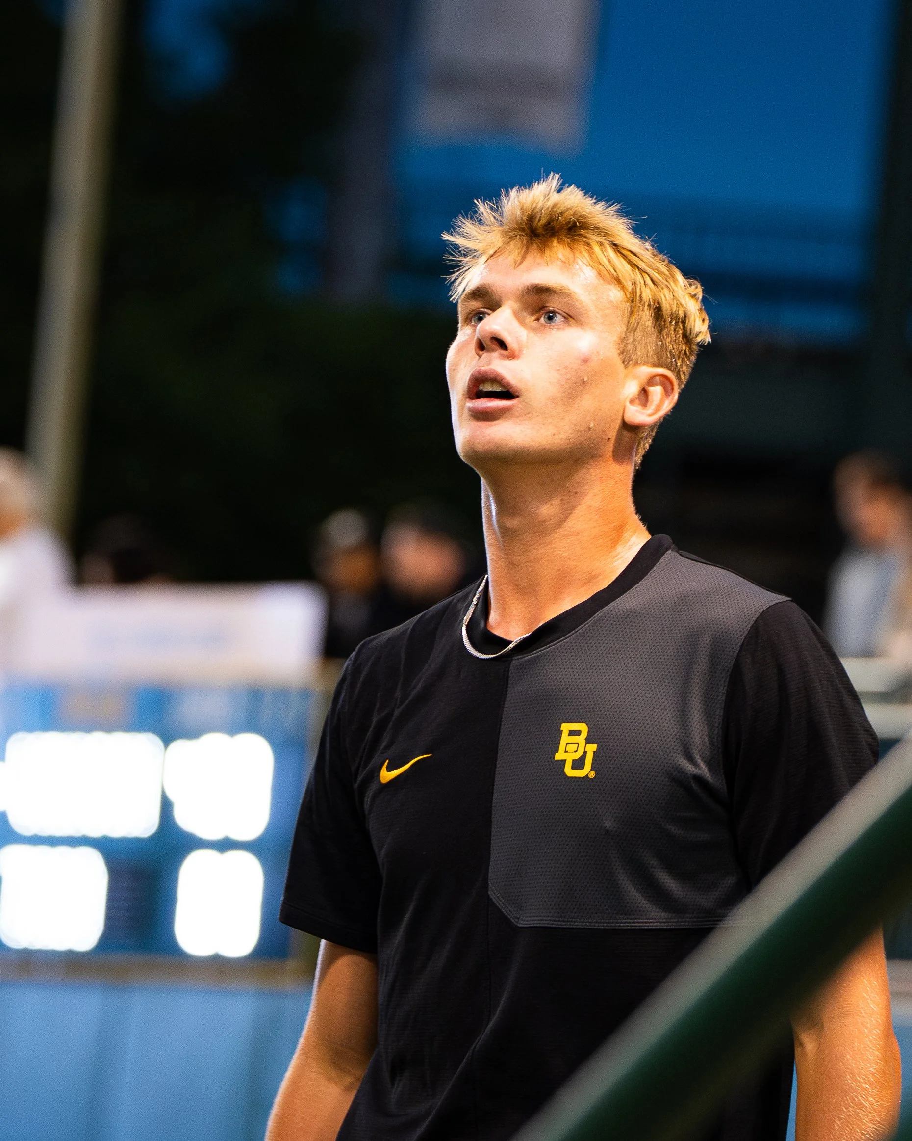 Young male tennis player wearing black shirt with yellow logo and Nike swoosh, focusing attentively during a match indoors.