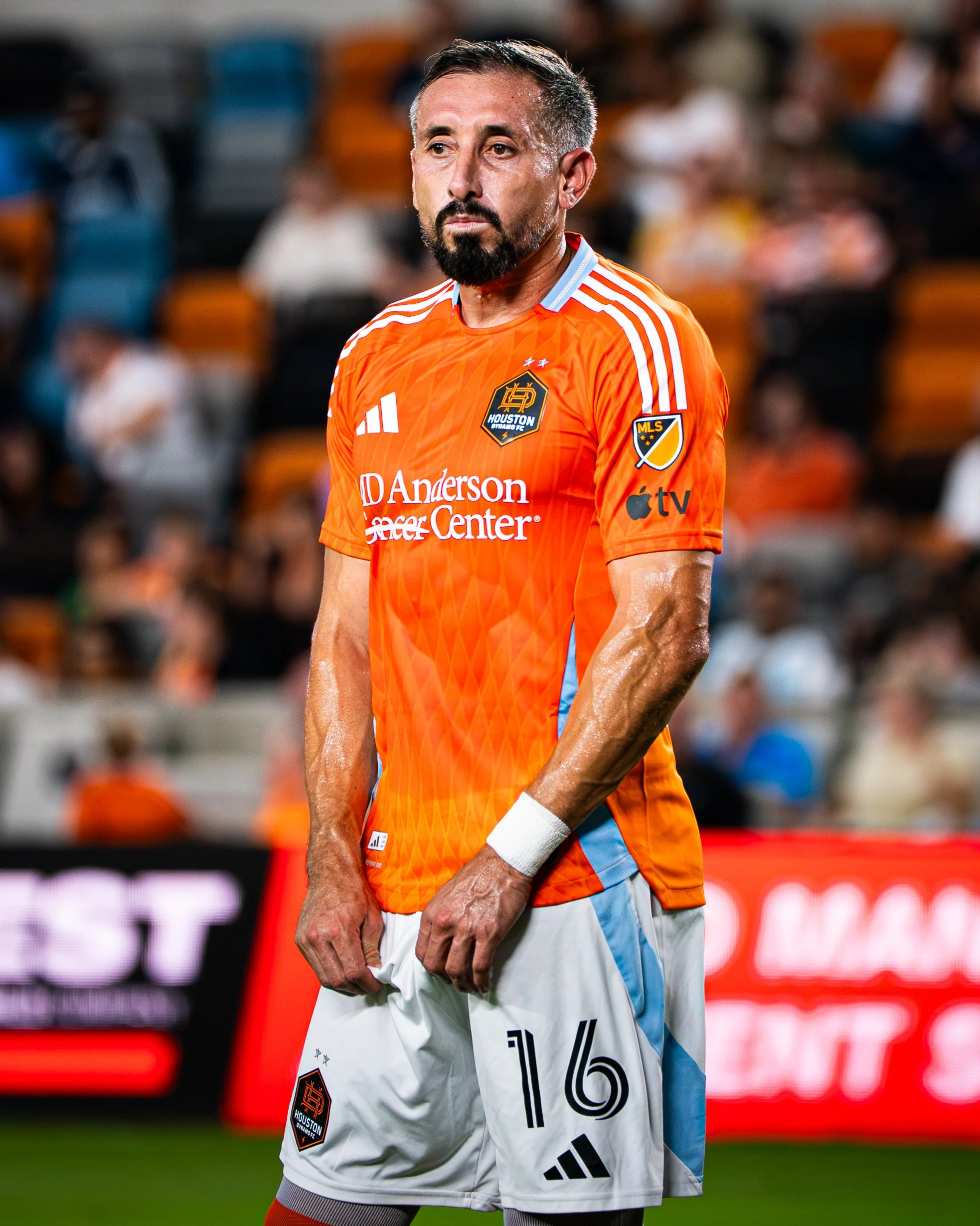A soccer player standing on the field wearing an orange Houston Dynamo FC jersey with the MLS logo on the sleeve and holding his shorts. The player has a serious expression, short dark hair, a beard, and sweat on his face. The blurred background show