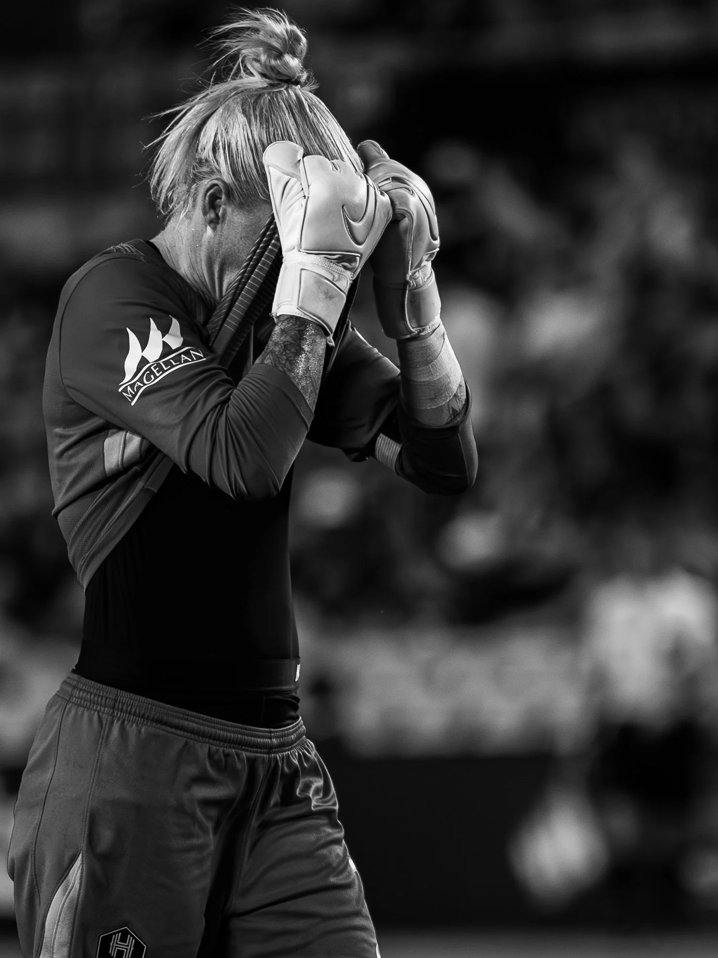 A baseball goalkeeper is praying with closed eyes, wearing gloves, a black sports shirt with a logo on the sleeve, and standing outdoors.