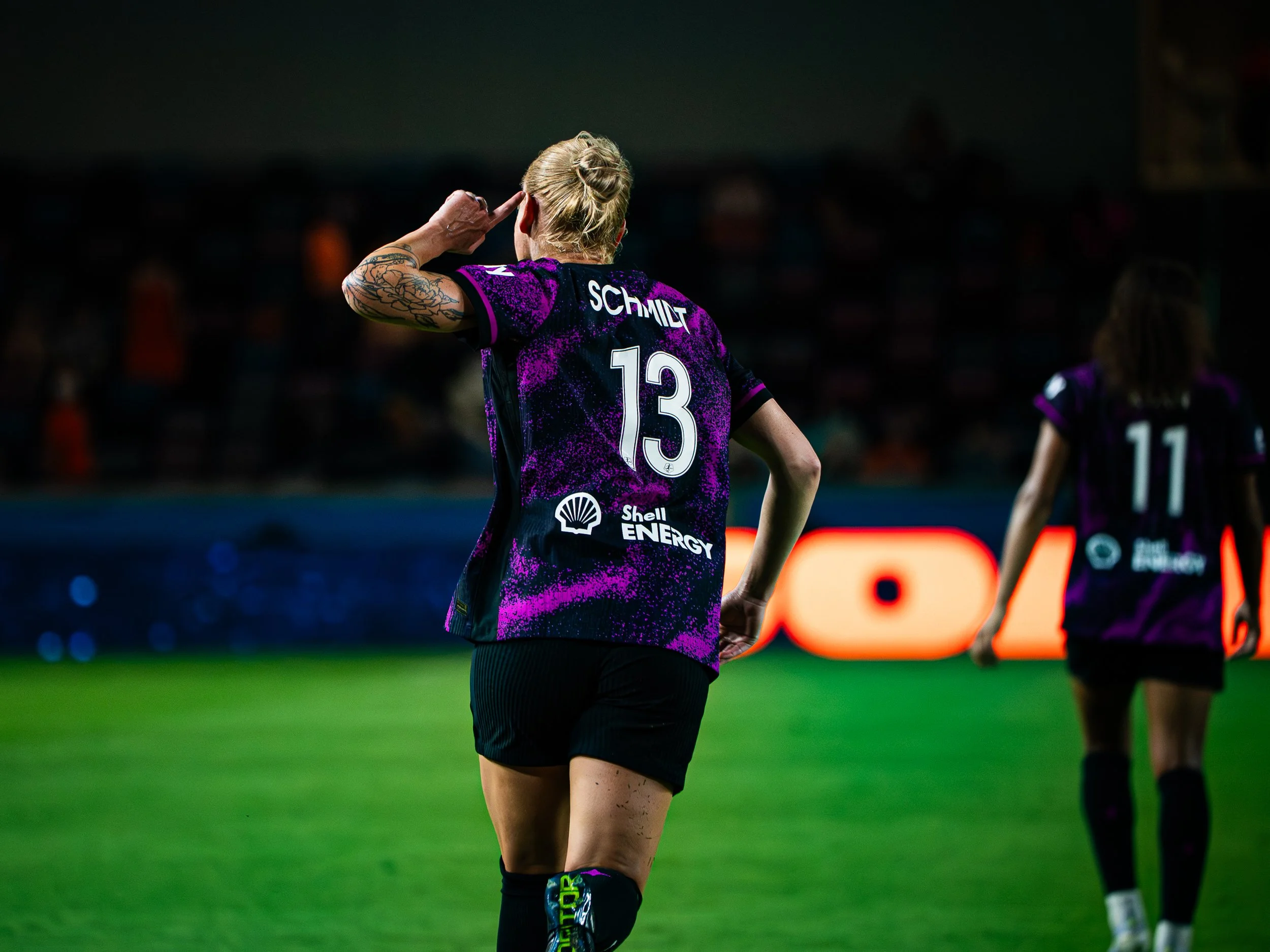 A female soccer player with blonde hair tied back, wearing a purple and black jersey with the number 13 and the name 'SCHMITT' on the back, is celebrating on a soccer field during a game at night.