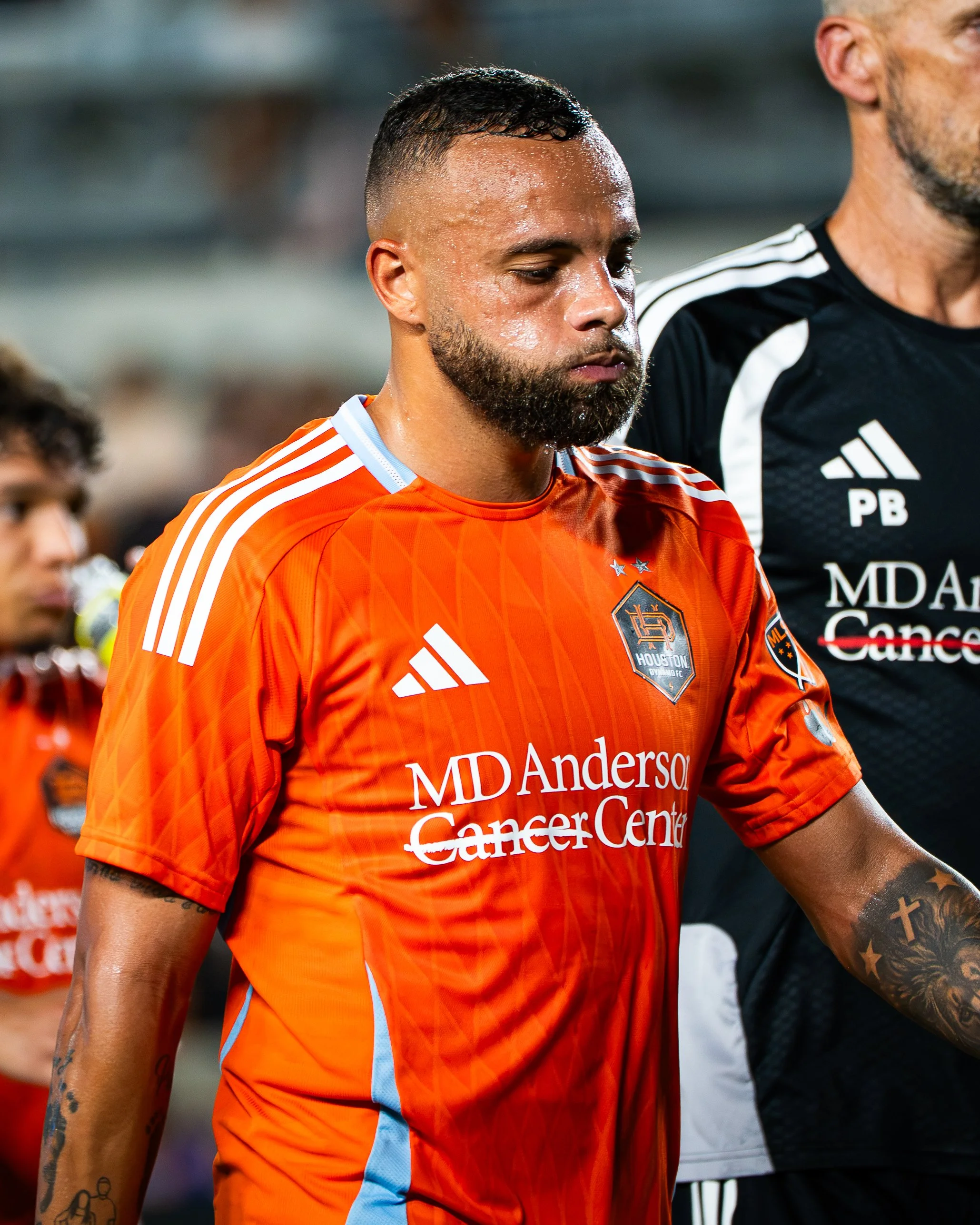 A man with a beard and short dark hair in a close-up shot, wearing an orange jersey with white stripes on the shoulders and logos for MD Anderson Cancer Center and Houston Dynamo FC. He appears to be in a sports setting, possibly after a game or duri