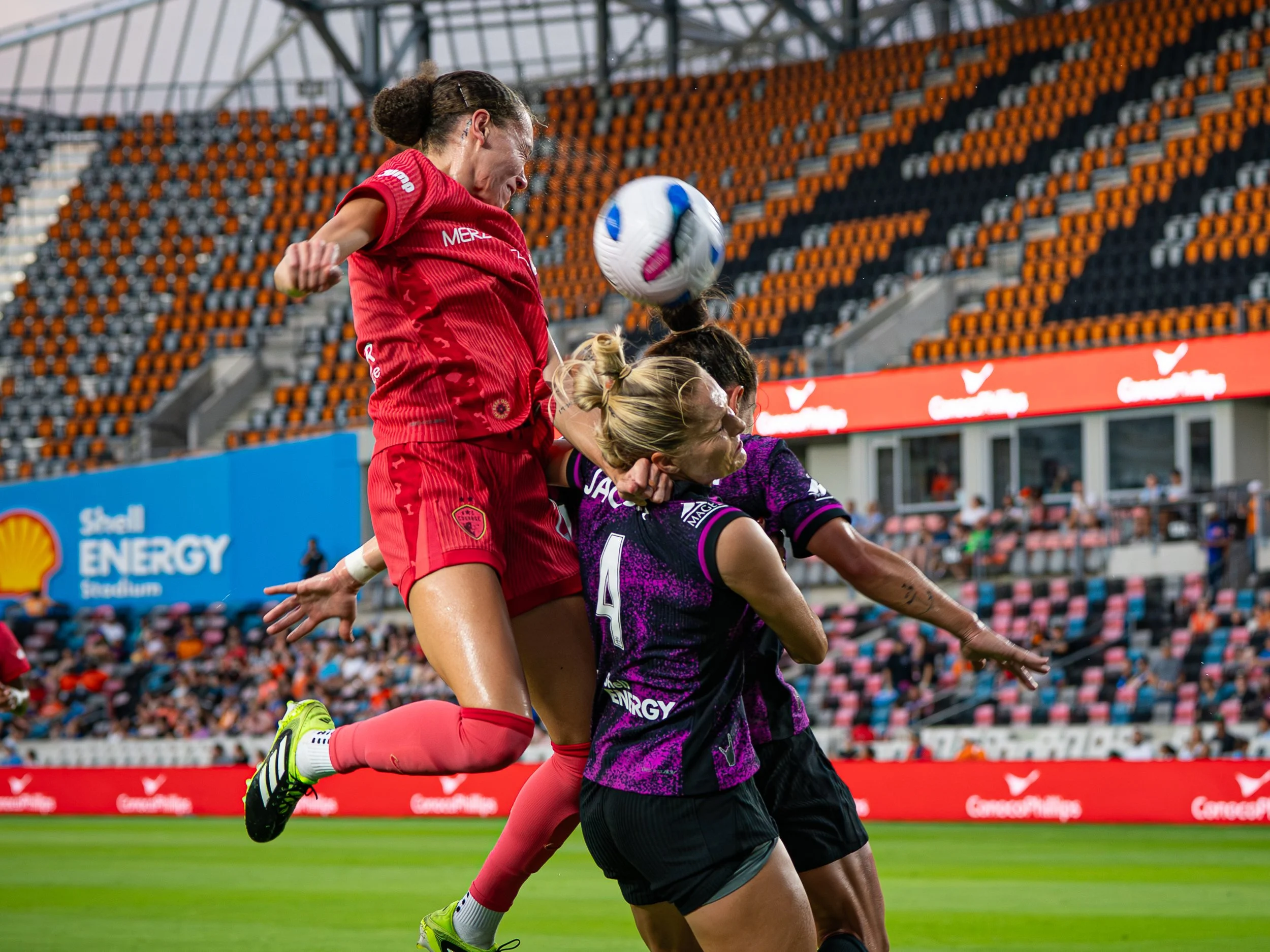 Two female soccer players jump and challenge for a header ball during a match in a stadium filled with spectators. One player is in a red uniform, and the other is in a purple and black uniform.