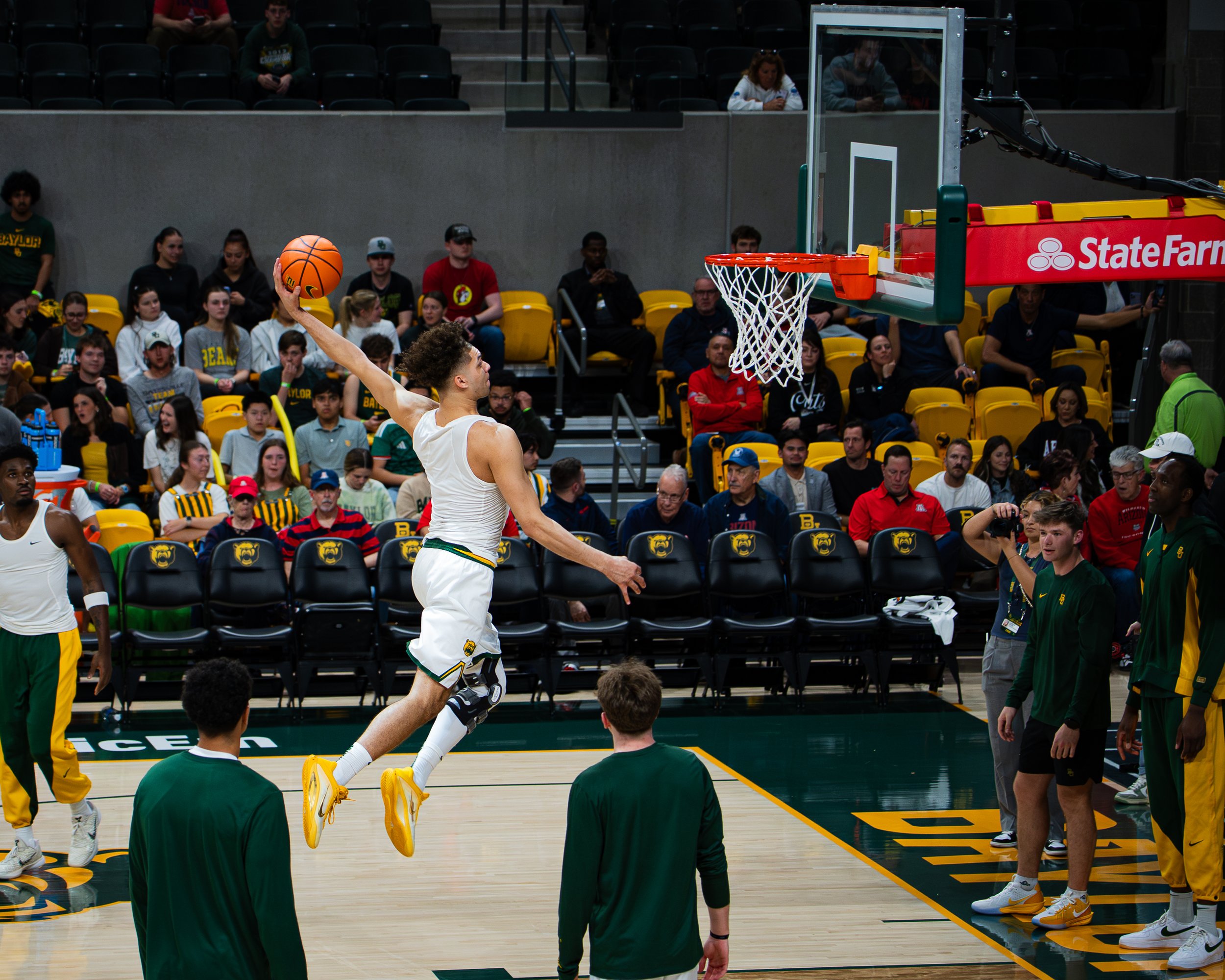 Basketball player in white uniform dunking the ball during a game, with an audience watching from behind.