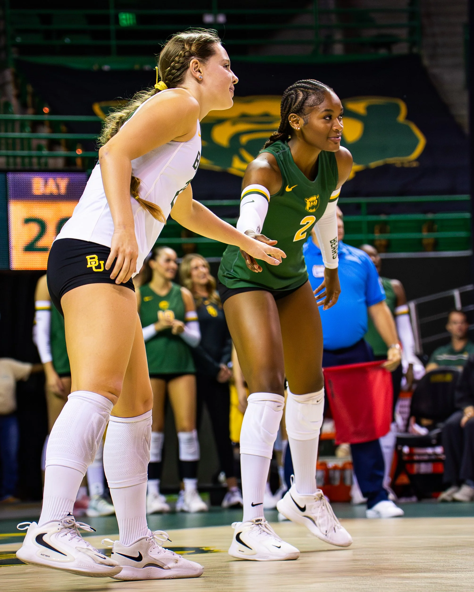 Two female volleyball players in white and green jerseys, standing in a ready position on the court with a blurred audience and other players in the background.