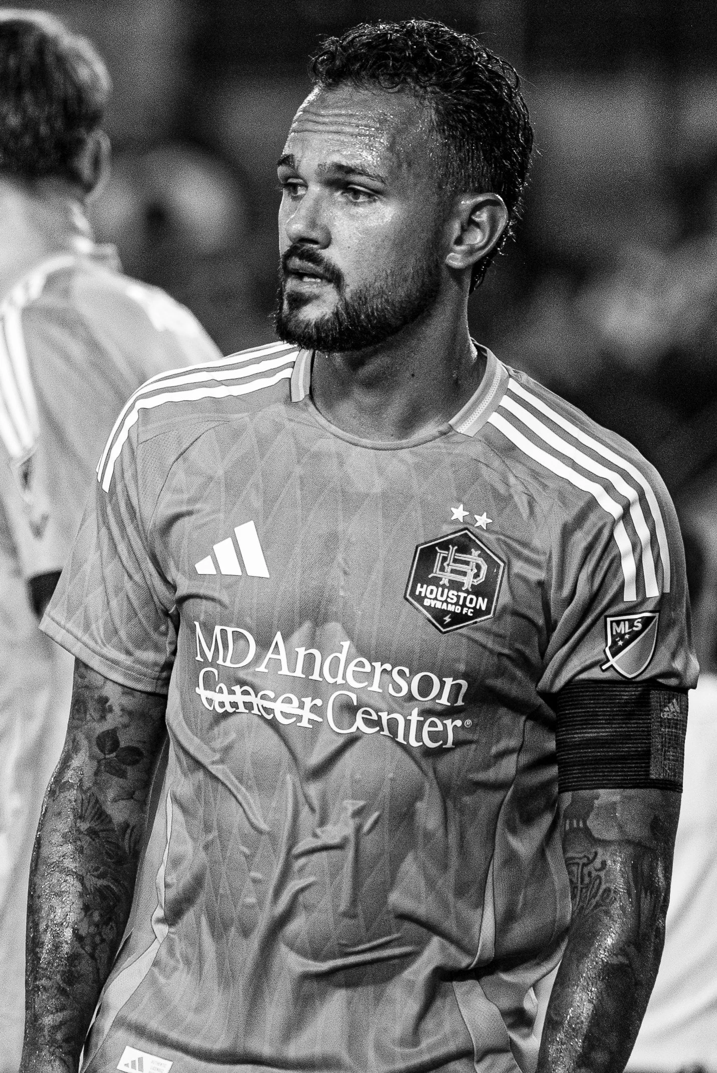Black and white photo of a male soccer player looking to his side, wearing a Houston Dynamo FC jersey with sponsor logo and MLS badge, with tattoos visible on his arms.