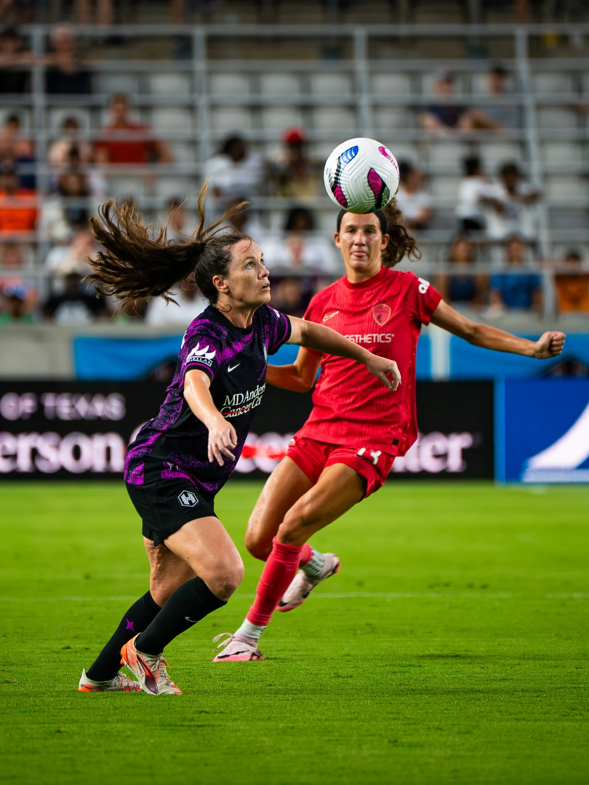 Two women playing soccer on a field, one in a purple and black uniform and the other in a red uniform, with a soccer ball in midair between them. Spectators are seated in the background.