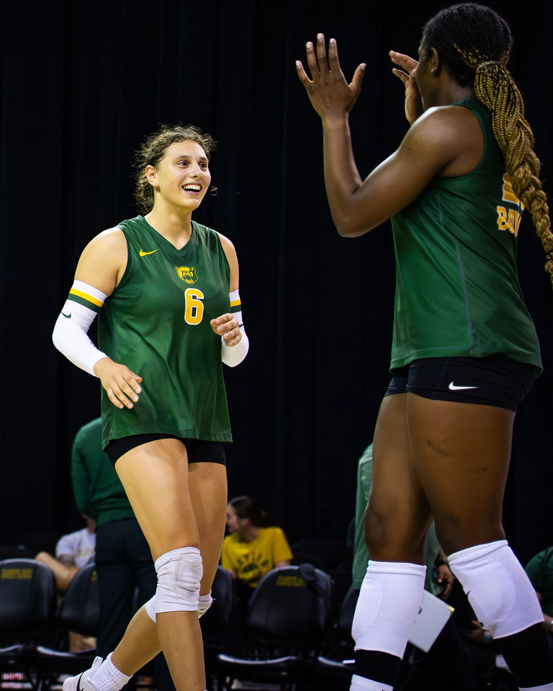 Two female volleyball players in green jerseys with yellow and white accents, one wearing number 6, are engaging in a conversation during a game or practice. They are indoors on a sports court, with a dark background and black chairs in the seating a