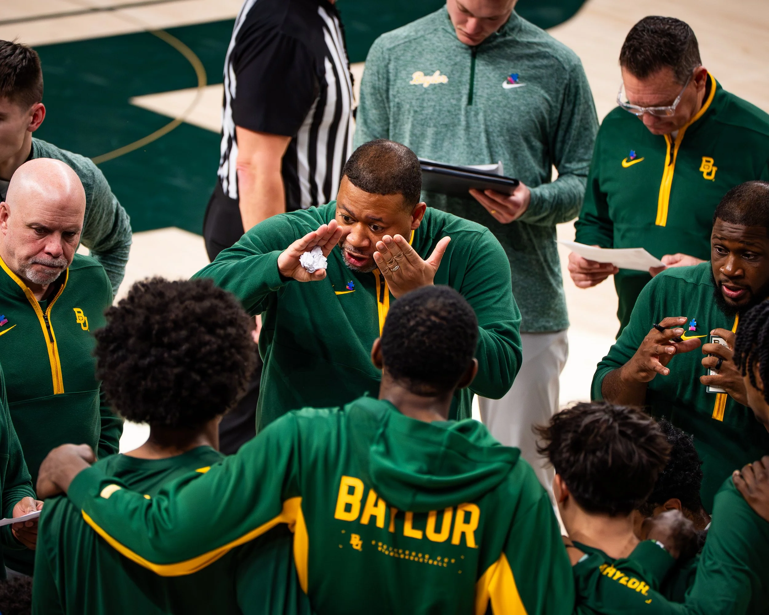 African American basketball coach talking to a team of young basketball players in green and yellow uniforms during a timeout on the court.
