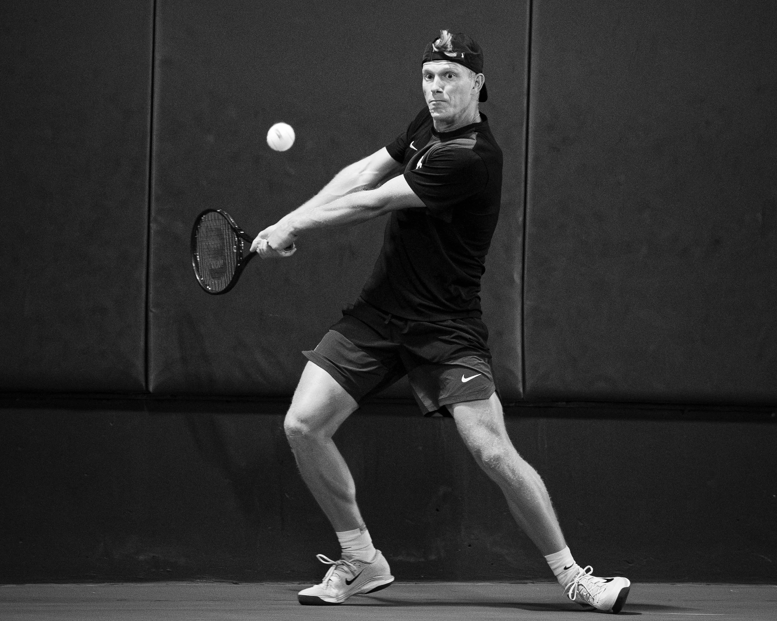 A man playing tennis indoors, preparing to hit the ball with his racket, wearing athletic clothing and a backward cap.