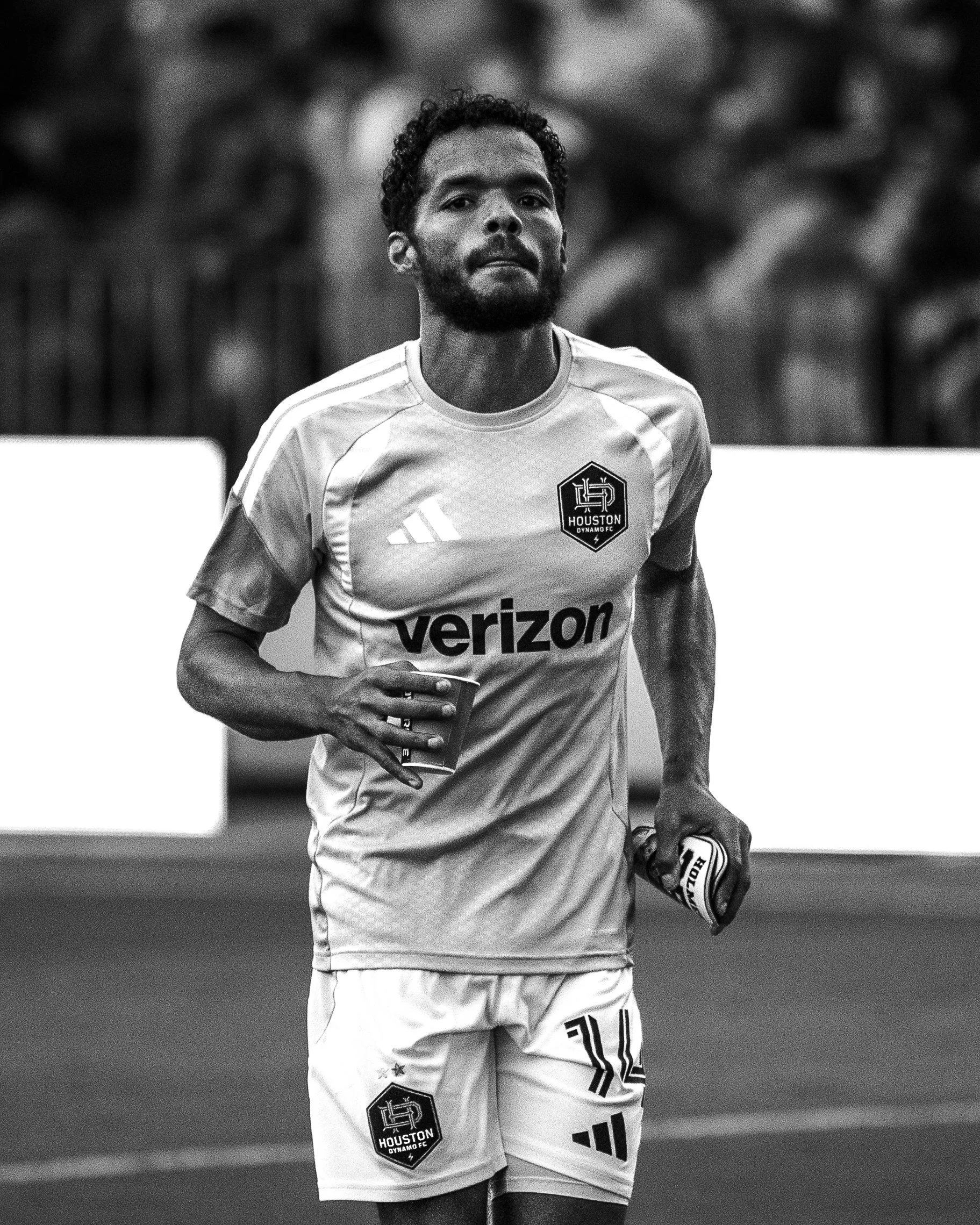 A male soccer player with dark curly hair and a beard, wearing a Houston Dynamo FC jersey, standing on a soccer field, holding a water bottle in his right hand and a soccer cleat in his left hand.