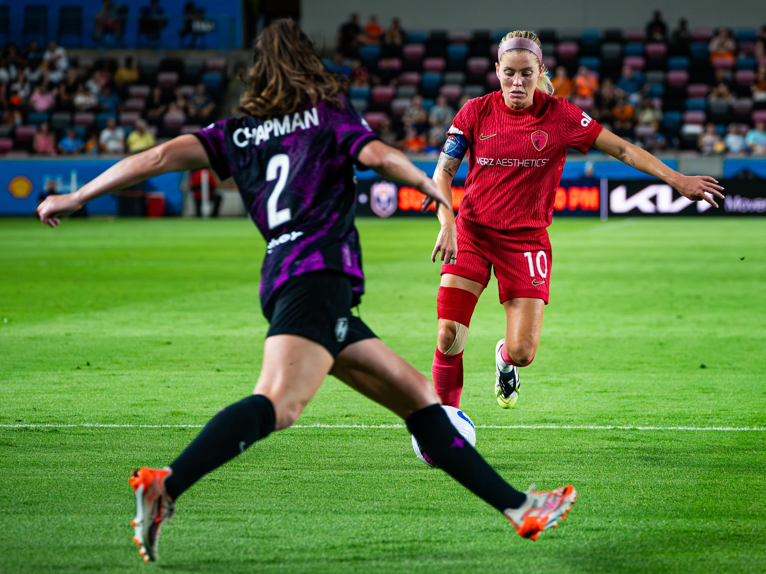 A soccer game with a female player in a red uniform controlling the ball while an opponent in purple attempts to block.
