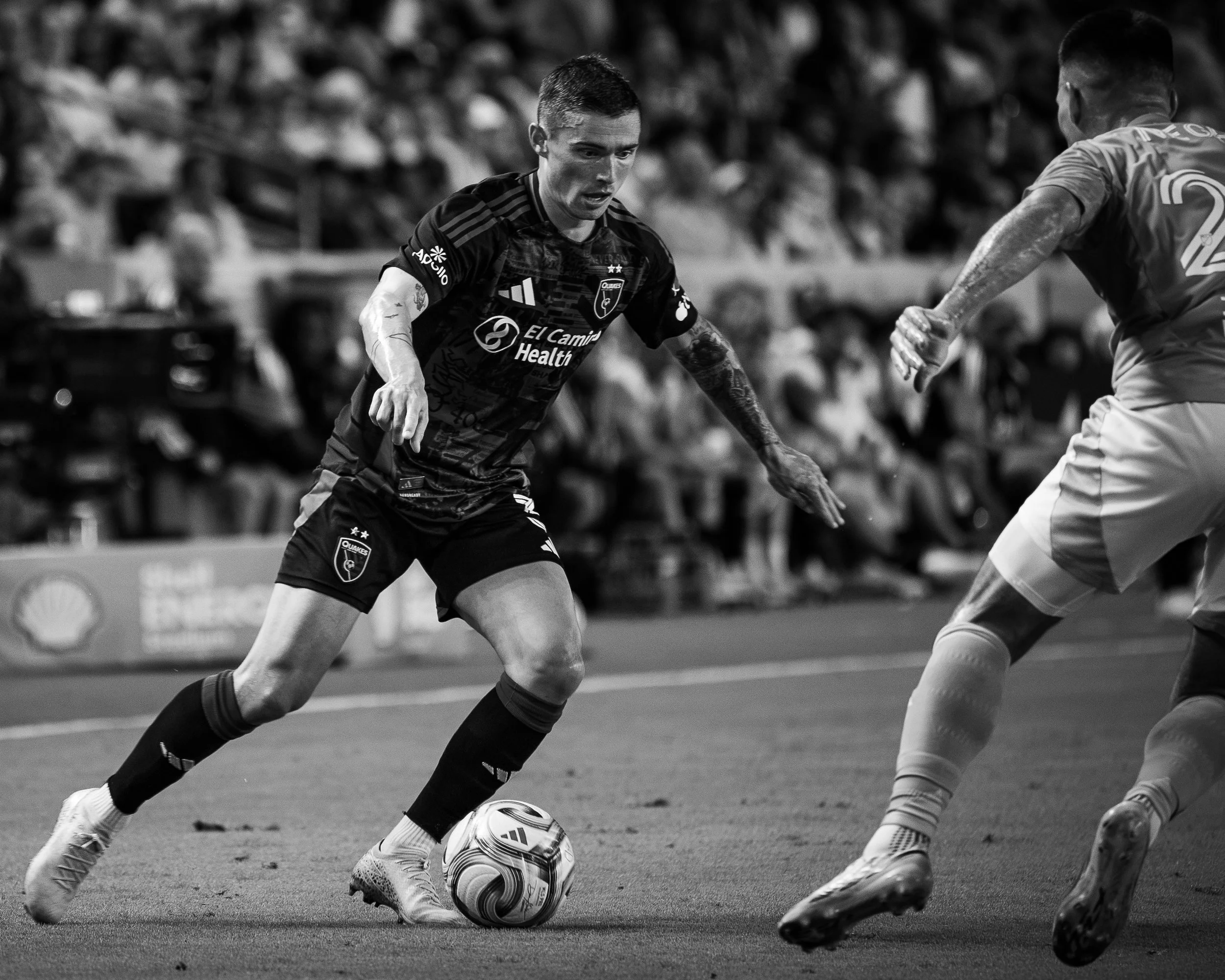 Black-and-white photo of two soccer players competing for the ball on the field, with a blurred crowd in the background.
