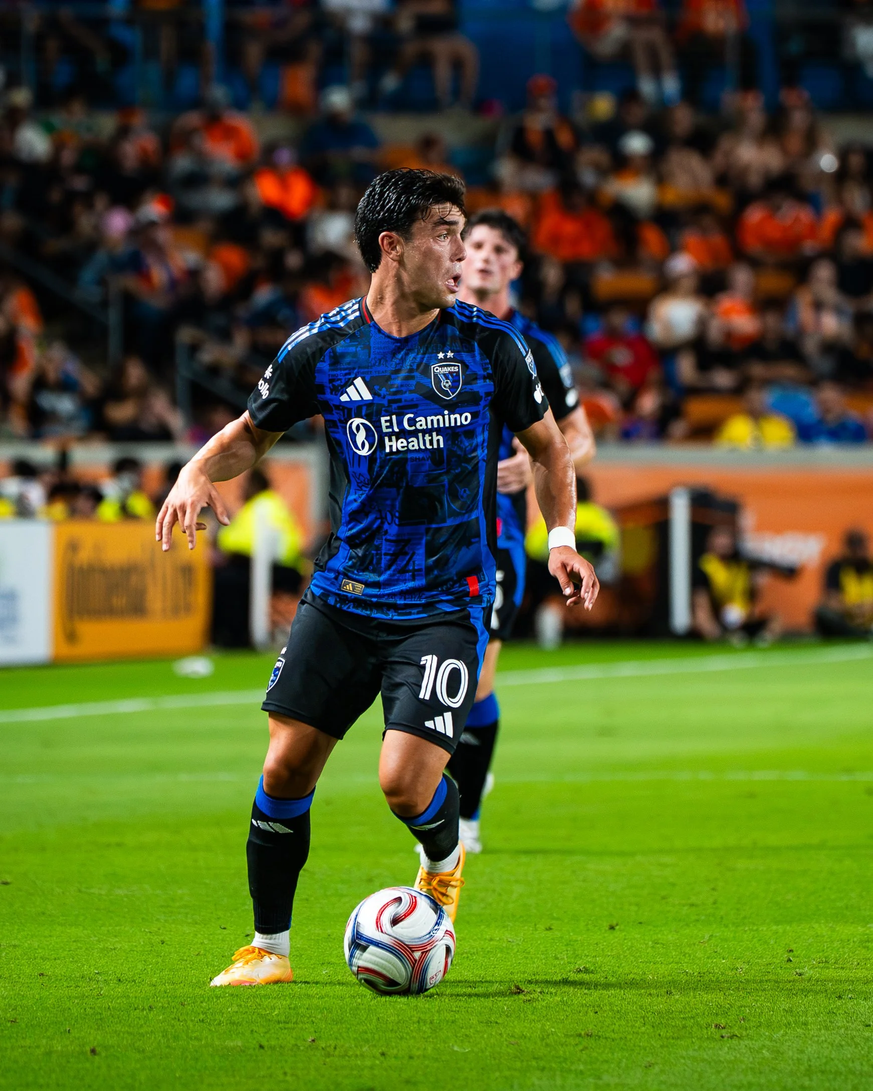 A soccer player in a blue and black uniform with the number 10 is about to kick a soccer ball on a green field, with a crowd of spectators in the background.