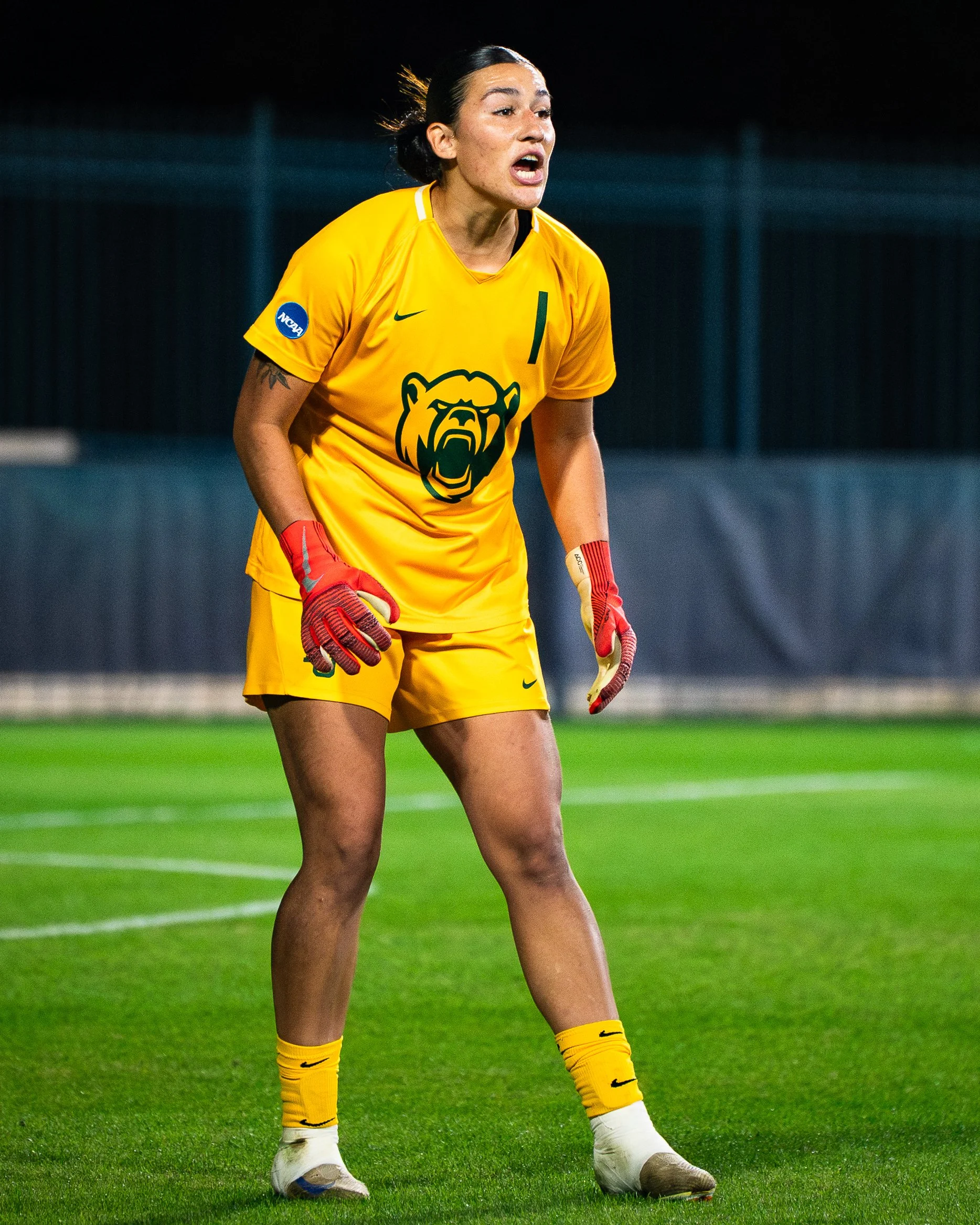 A female soccer player in a yellow uniform with team logo and gloves, standing on a green soccer field during a night game, appearing to shout.