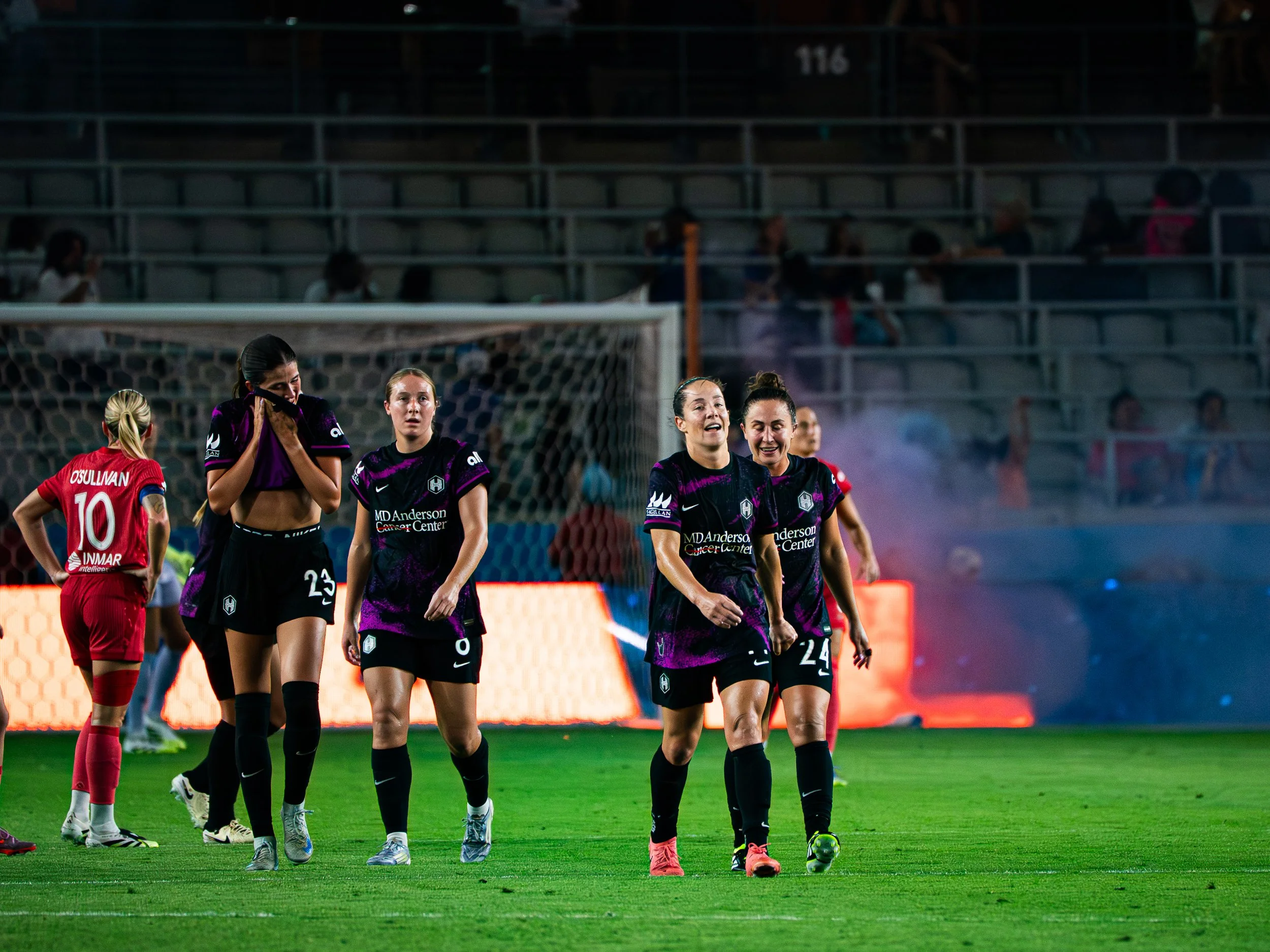 Women soccer players walking on the field during a game, with a stadium in the background.