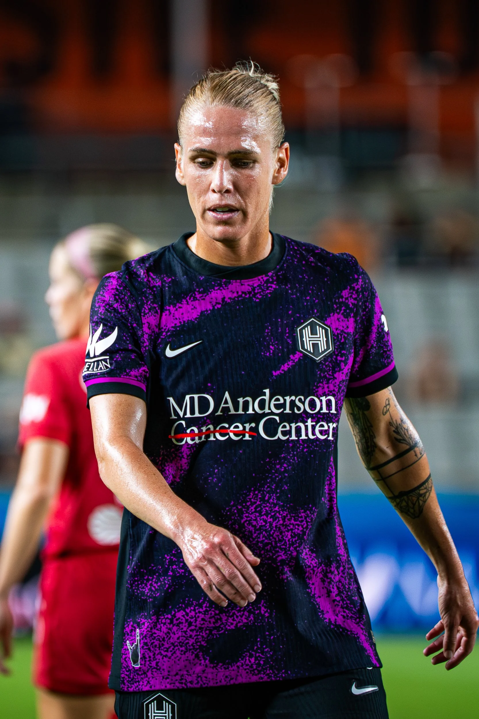 Female soccer player with blonde hair in a ponytail, wearing a purple and black jersey with the MDAnderson Cancer Center logo, walking on the field with a focused expression.