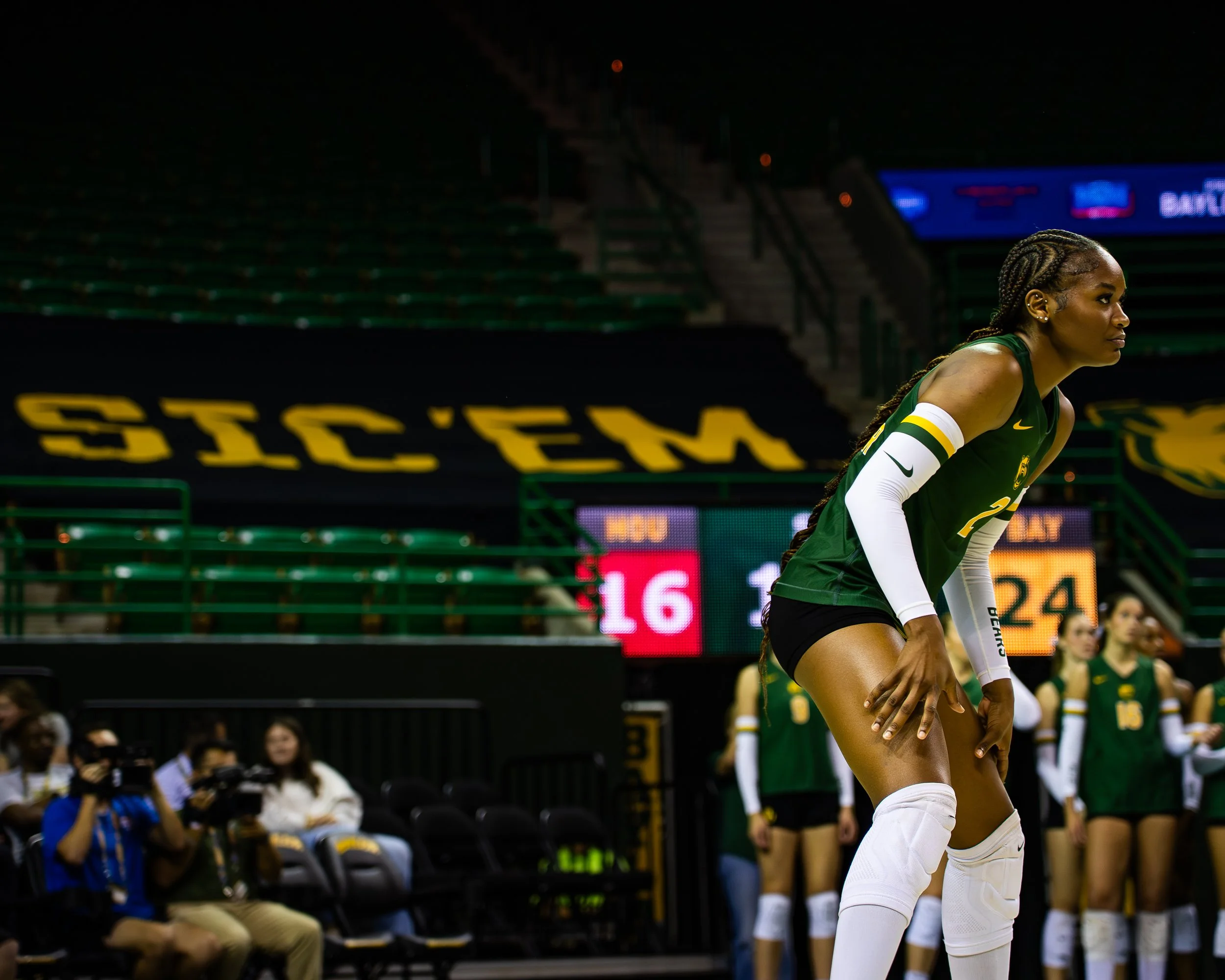 An athlete in a green and white uniform bent slightly forward on a volleyball court, with her hands on her knees, at an indoor stadium during a game or practice session.