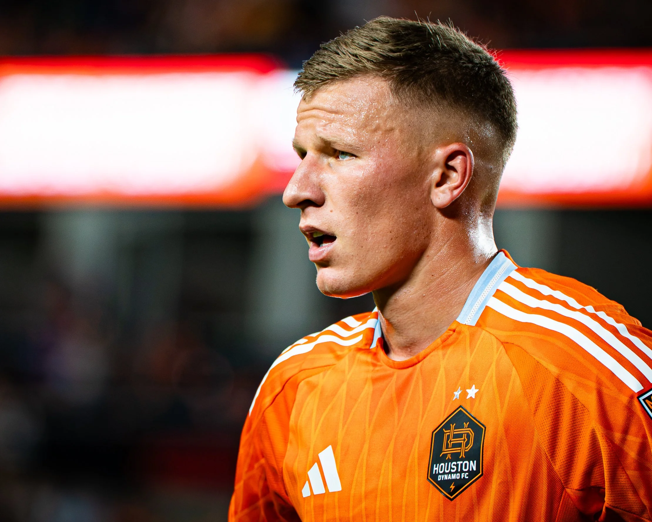 Close-up of a male soccer player wearing an orange Houston Dynamo FC jersey, showing part of his face and upper chest, with a background of blurred lights.