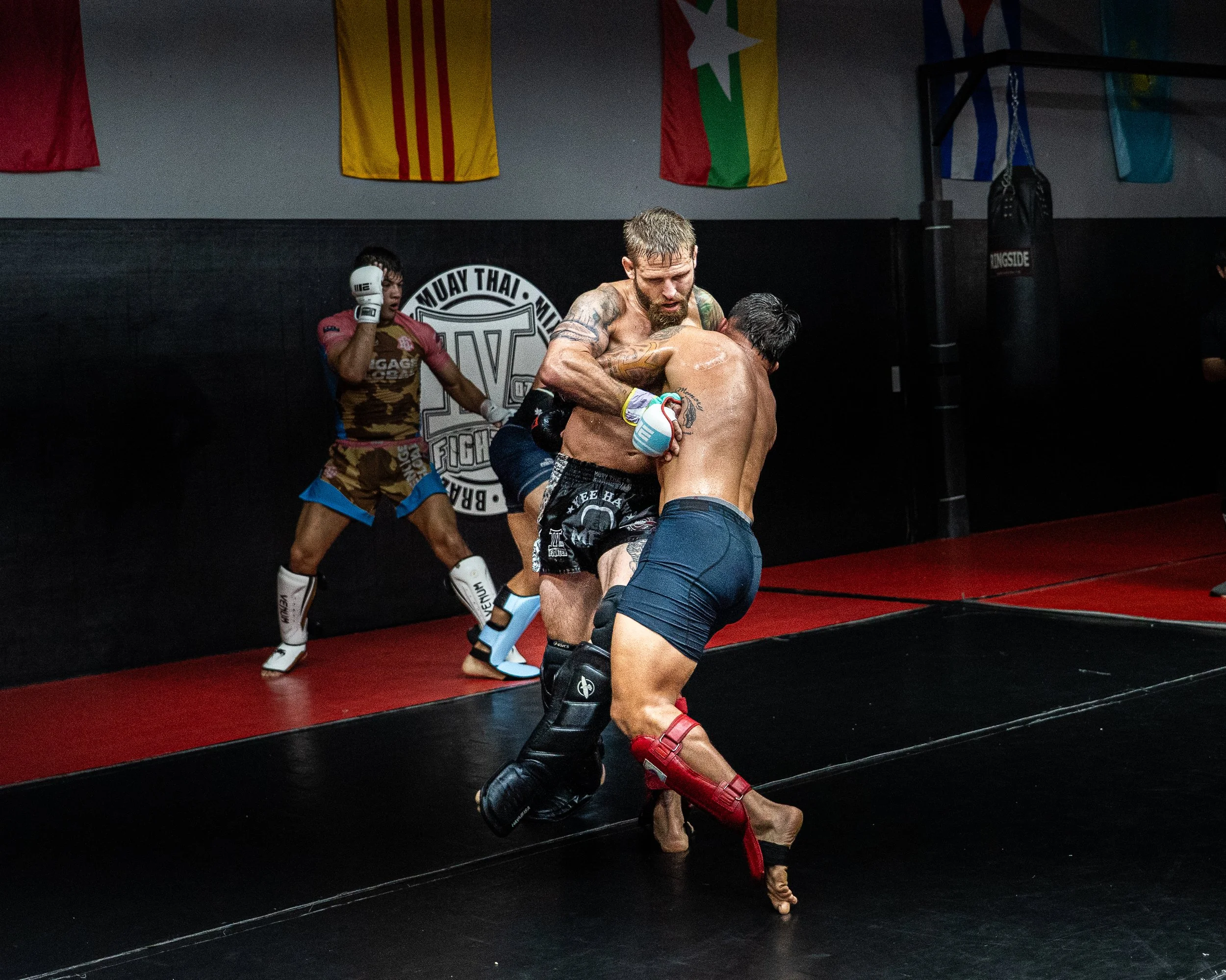 Two Muay Thai fighters sparring in a gym with flags hanging on the wall behind them. One fighter has a beard and tattoos, the other is younger with dark hair. A female fighter in sportswear stands in the background, watching the match.