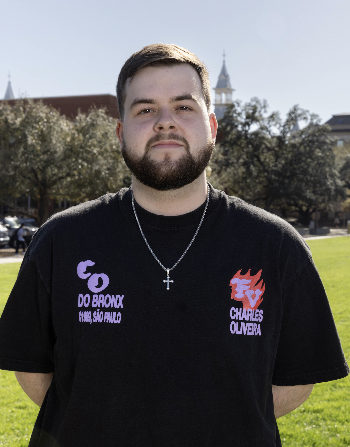 A young man with a beard and short hair standing outdoors in front of trees and a historic building. He is wearing a black T-shirt with purple and red text, and a silver chain with a cross pendant.