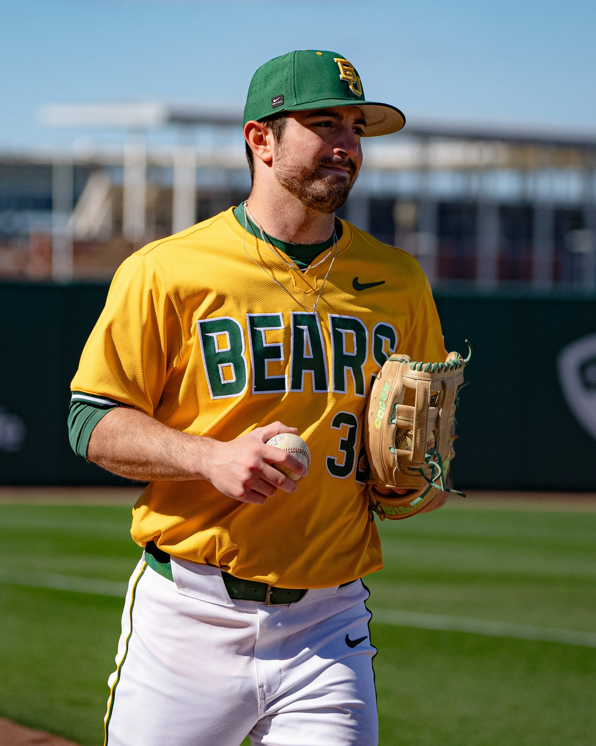 A man wearing a green baseball cap, yellow jersey with 'BEARS' on it, and white baseball pants holding a baseball during daytime on a baseball field.